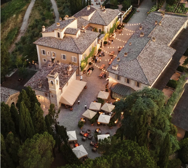 Aerial view of a courtyard with outdoor dining, string lights, and umbrellas surrounded by Mediterranean-style buildings with tiled roofs, lush greenery, and trees. Tuscany Rosewood castiglon de Bosco