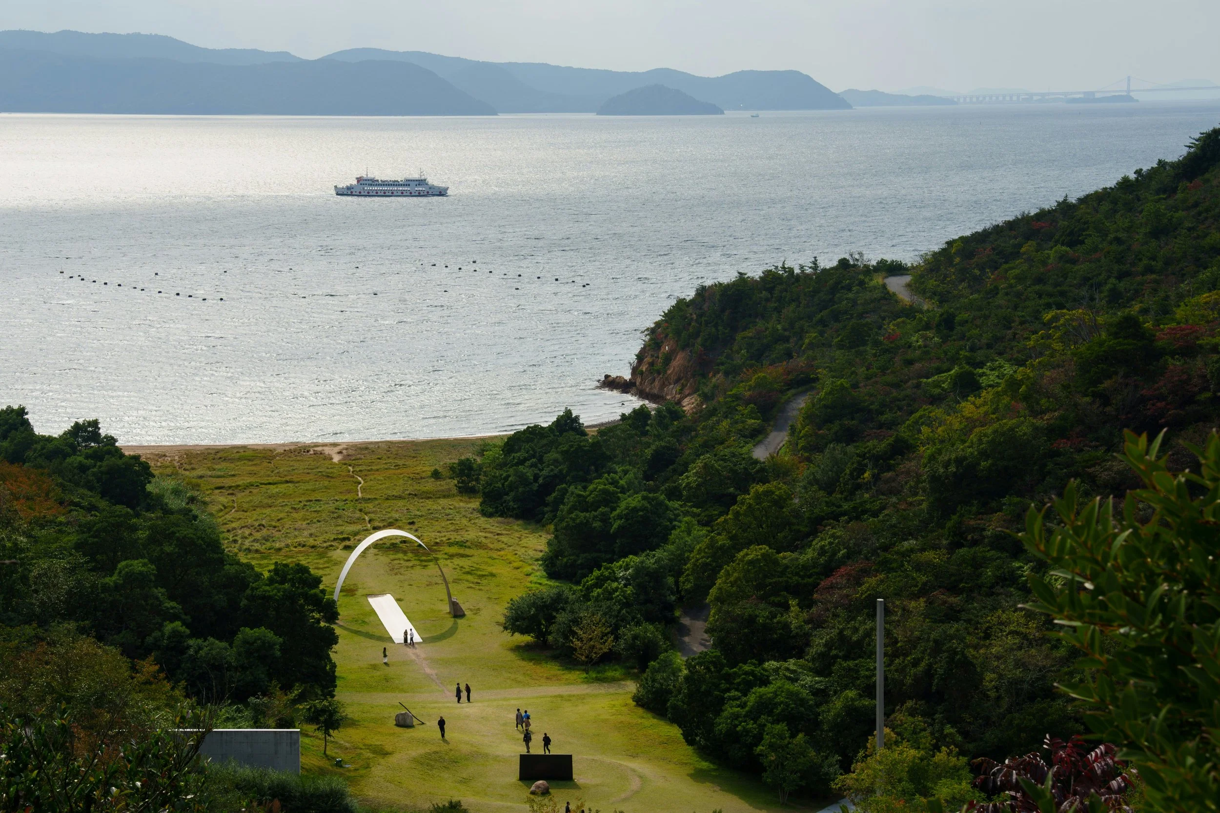A green valley on Naoshima leading down to the Seto Inland Sea, with a white arch sculpture on the lawn and a ferry crossing the water towards distant islands.