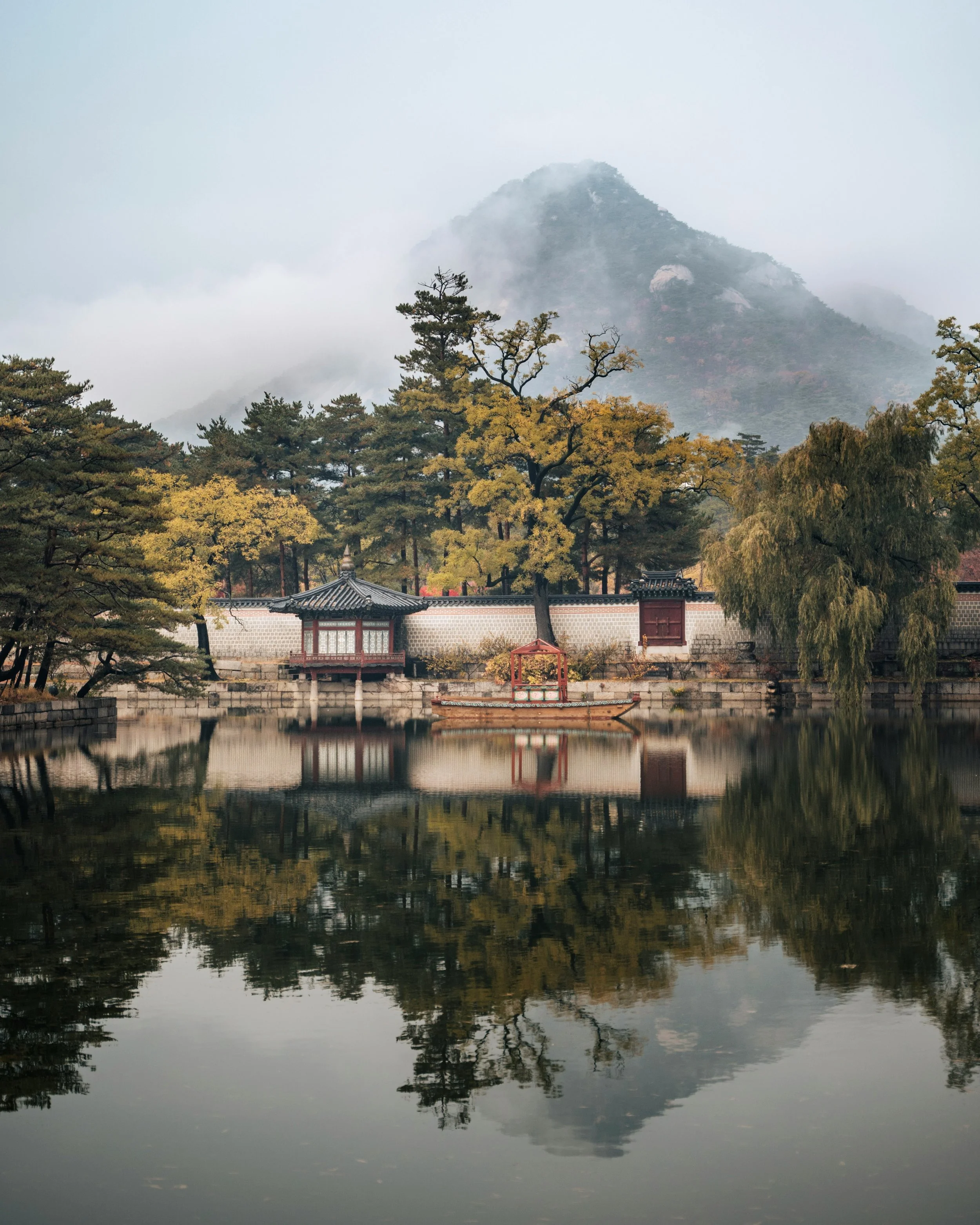 Mountains shrouded in fog with trees in the foreground and a traditional building by the water. Seoul palace tour