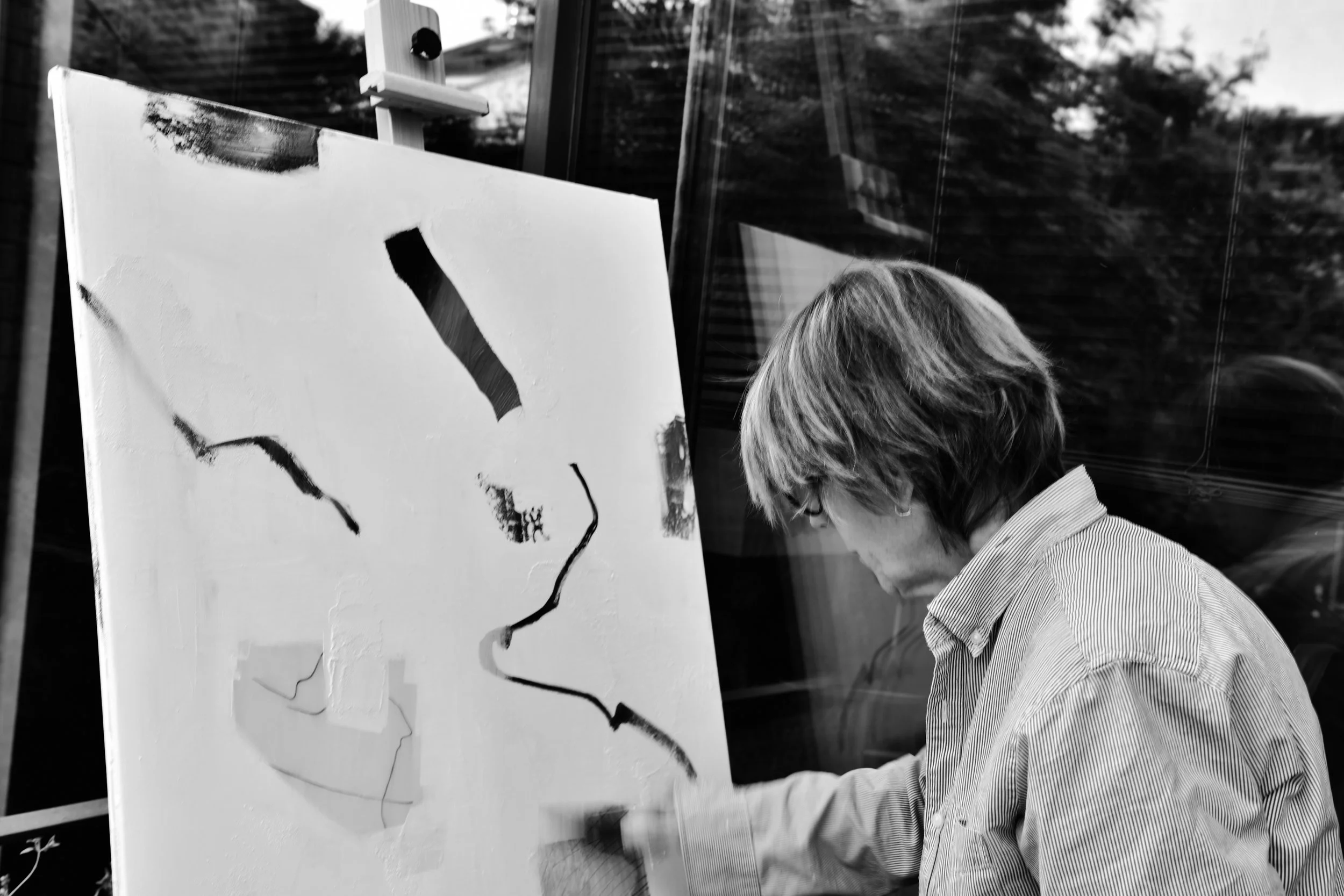 A person with short hair and glasses painting a portrait on a canvas outdoors, with trees visible in the background.
