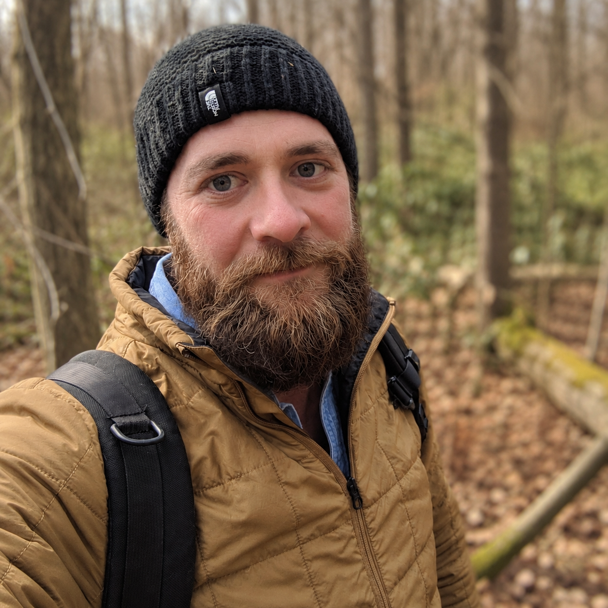 A bearded man in a black beanie and tan jacket takes a selfie in a forest with trees and fallen leaves in the background.