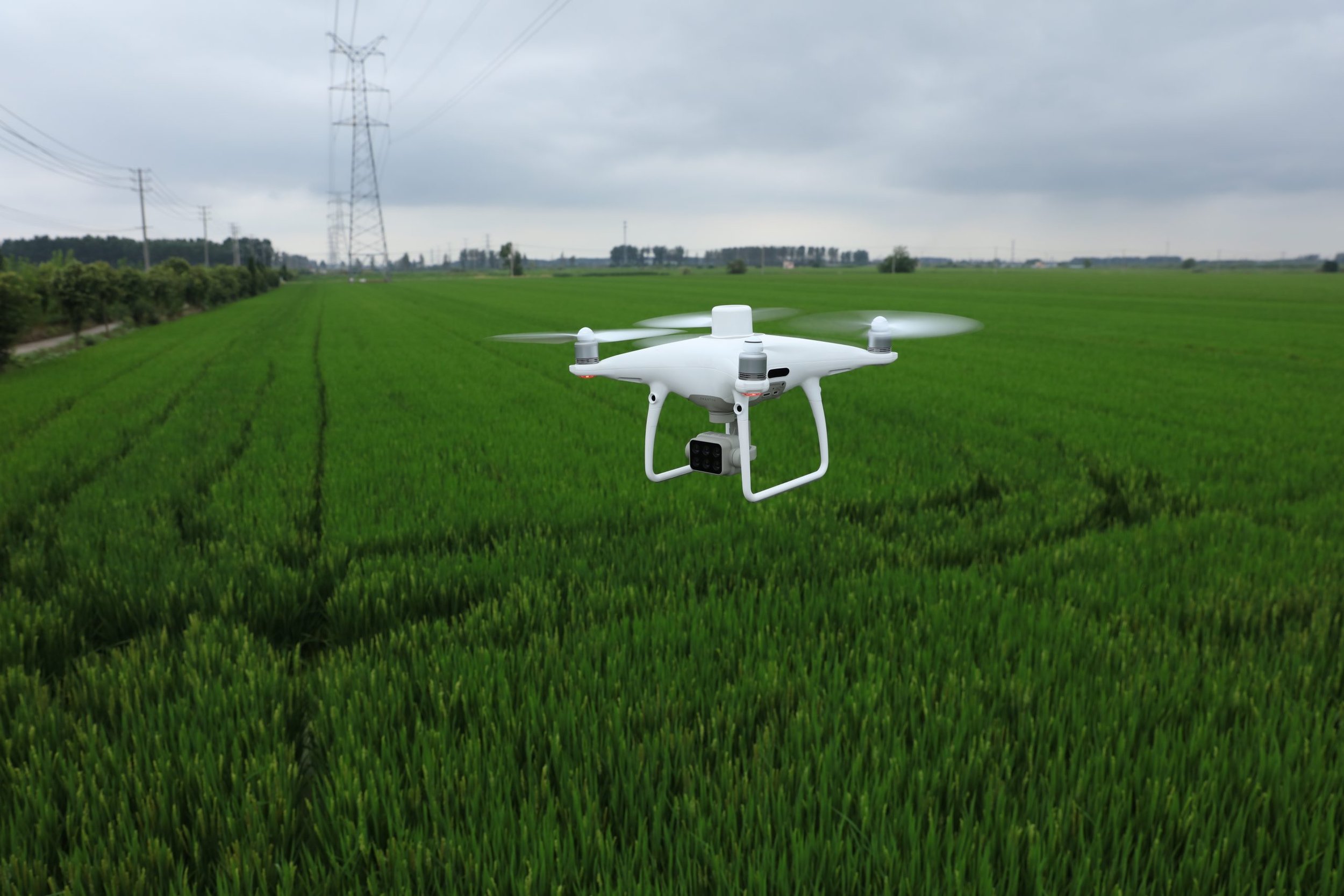 A drone flying over a green agricultural field on a cloudy day, with power lines and transmission towers in the background.