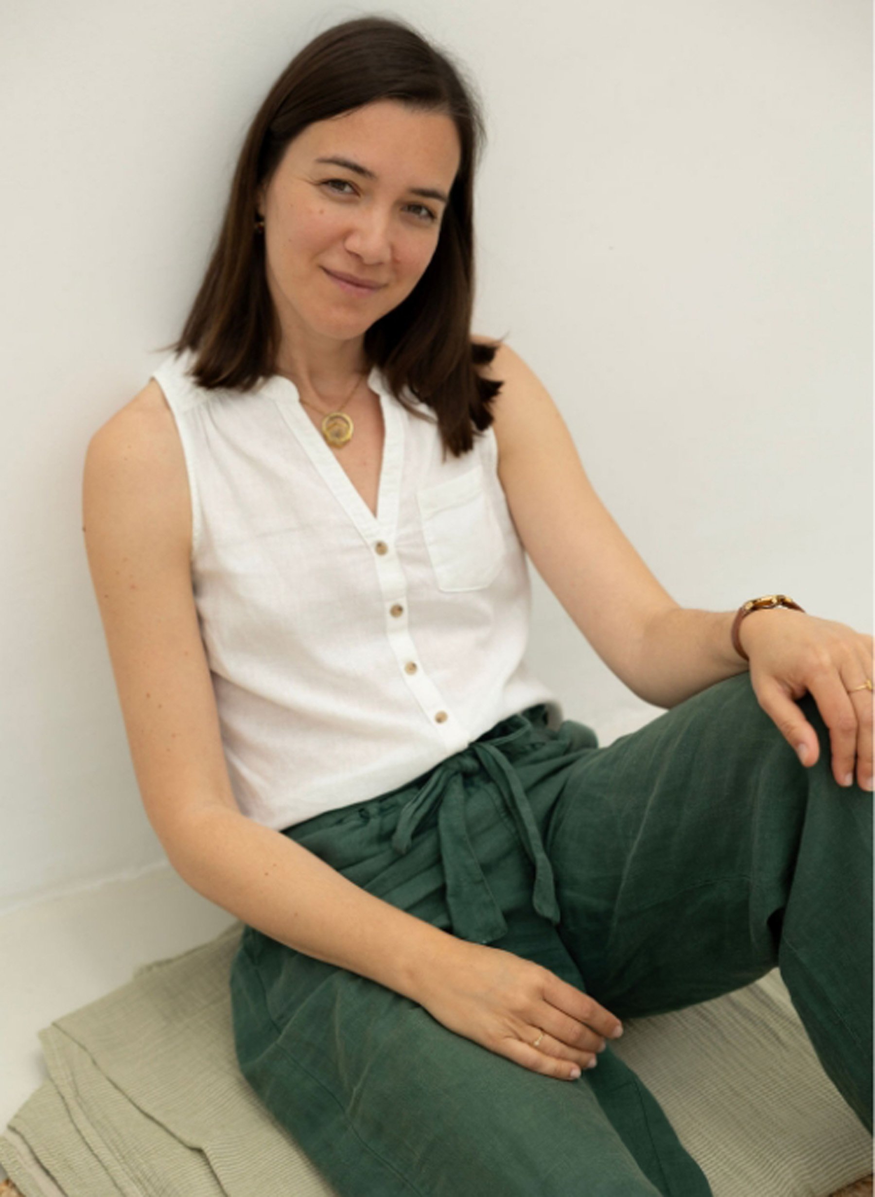 A woman with brown hair, wearing a sleeveless white blouse with buttons, dark green pants, and a gold necklace, sitting on a beige mat against a plain white wall.
