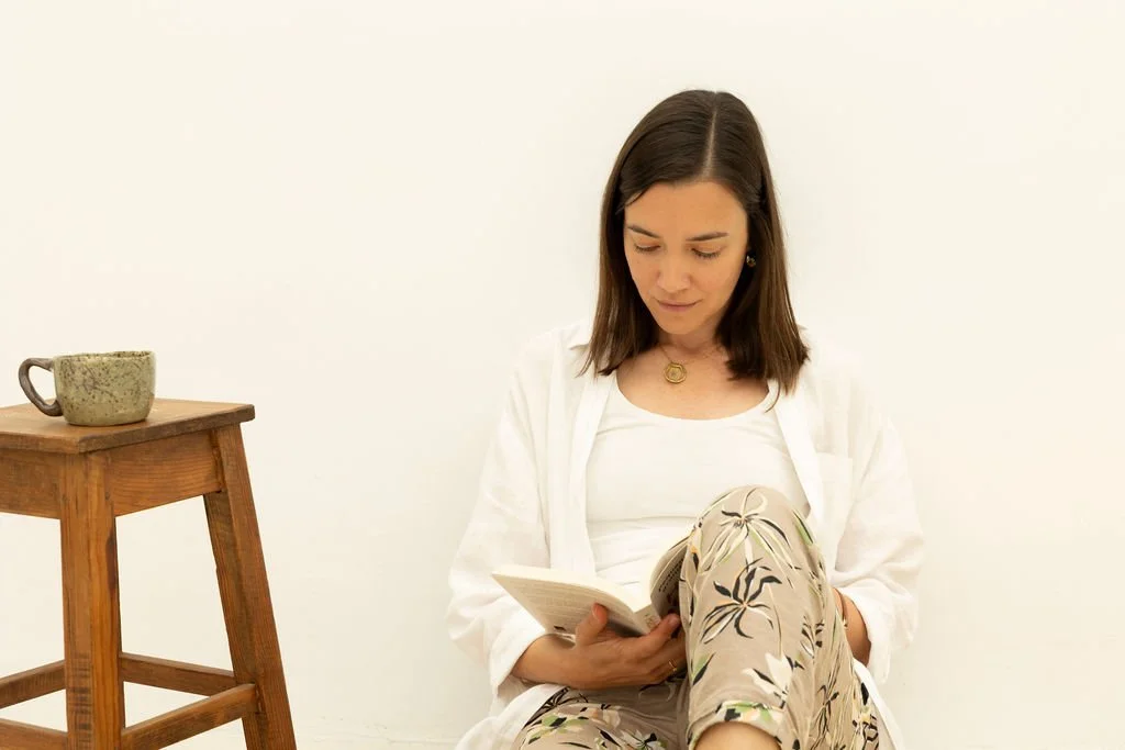 Woman sitting on the floor reading a book, next to a wooden table with a mug on it, against a plain white wall.