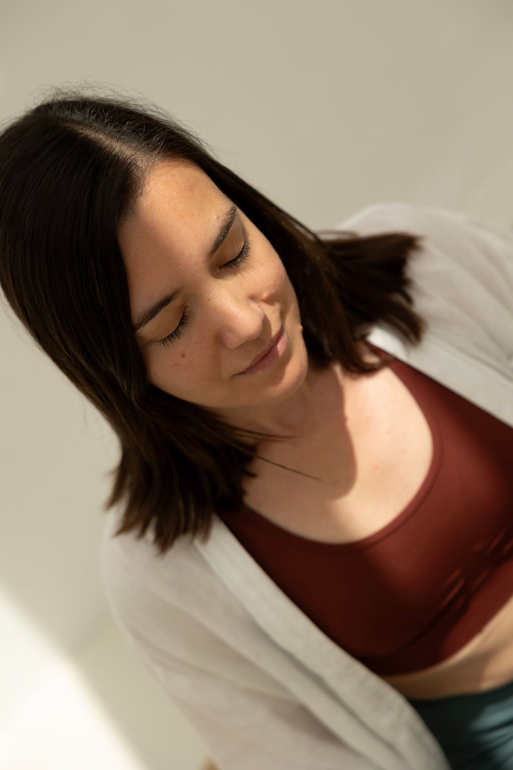 A young woman with shoulder-length brown hair, closed eyes, and a slight smile, wearing a red top and a white cardigan, standing against a plain light-colored background.