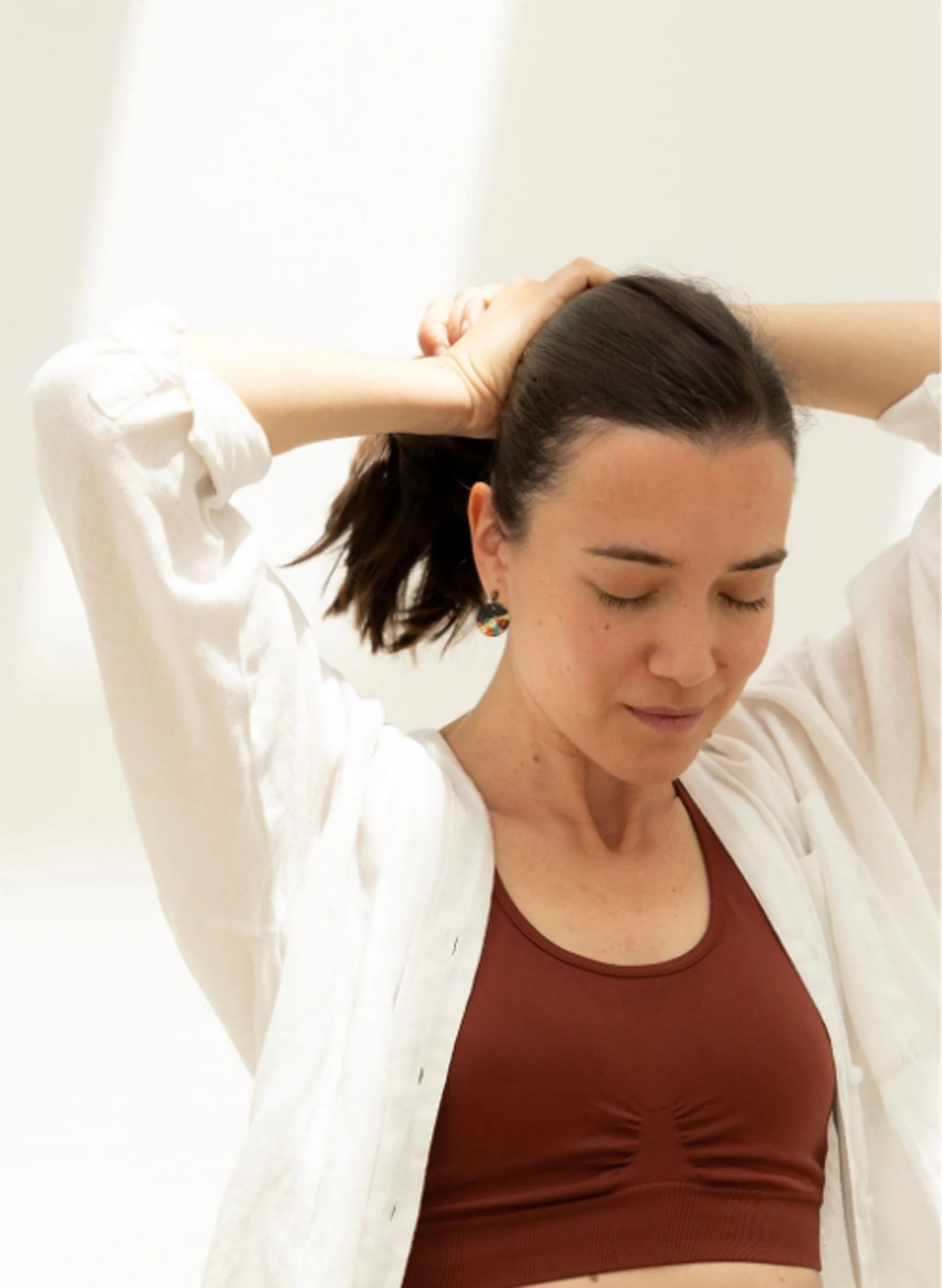 A woman with dark hair, wearing a rust-colored top and a white shirt, stands against a plain white background. She has her eyes closed and is touching her hair with both hands.