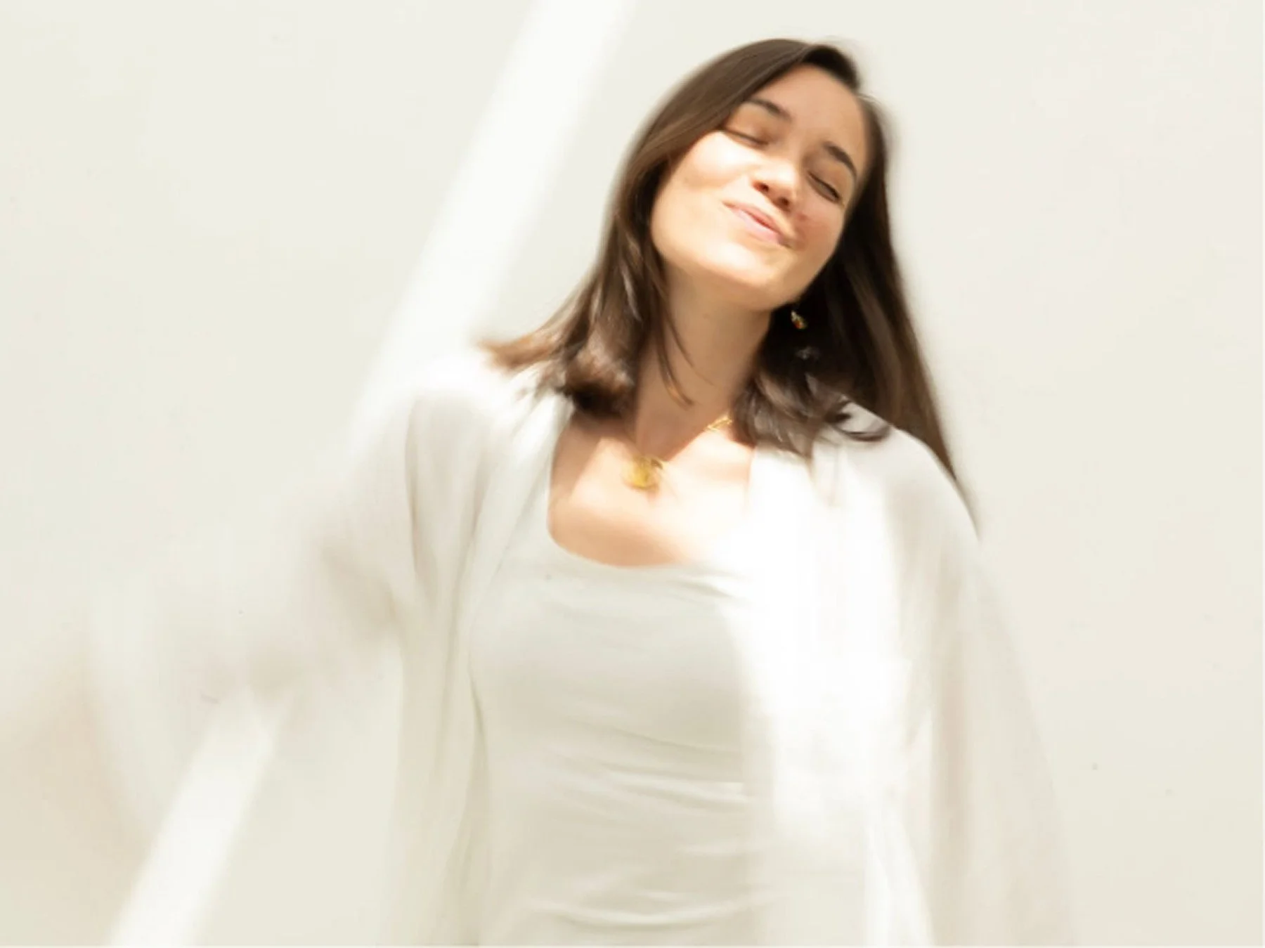 A woman with shoulder-length brown hair smiling with closed eyes, wearing a white top and gold jewelry, against a plain light-colored background.
