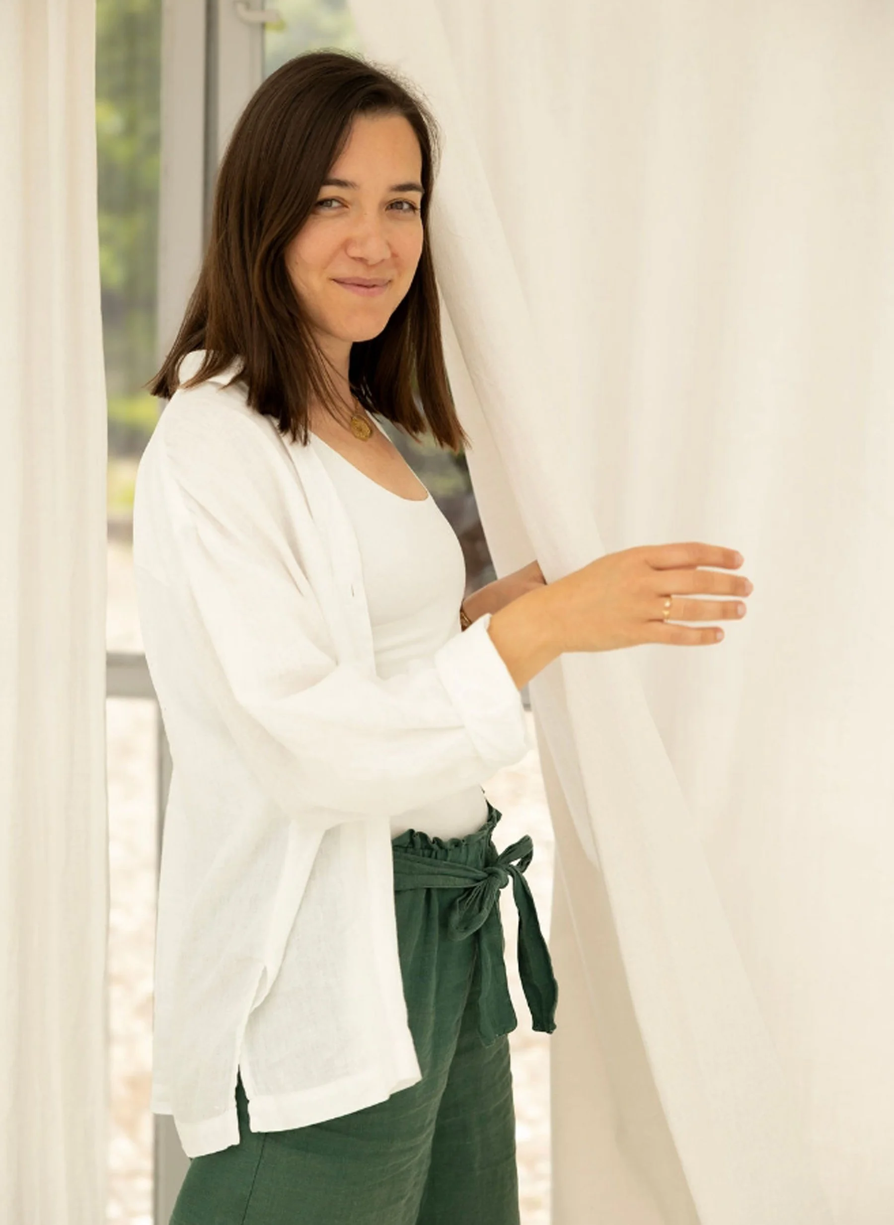 A woman with brown hair and light skin peeks out from behind white curtains, smiling softly while holding the curtains open. She is dressed in a white top and dark green pants with a matching tie, and is wearing a gold necklace. The background shows a window and natural light.