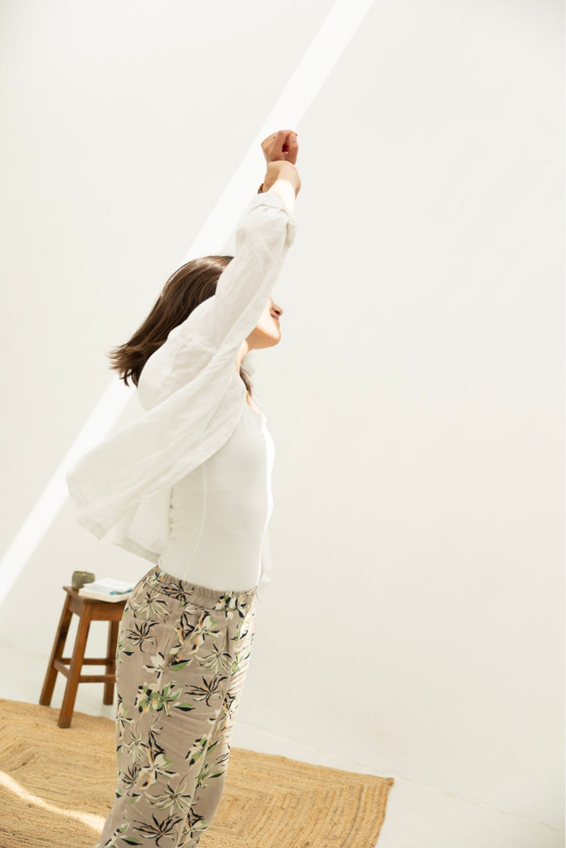 A woman standing in a room with her arm raised, holding a white pole, wearing a light-colored blouse and patterned pants with a floral design.