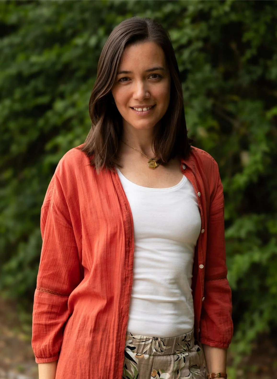 A young woman with shoulder-length brown hair wearing a white top, a red-orange button-up shirt, and patterned pants, standing outdoors in front of green leafy background.