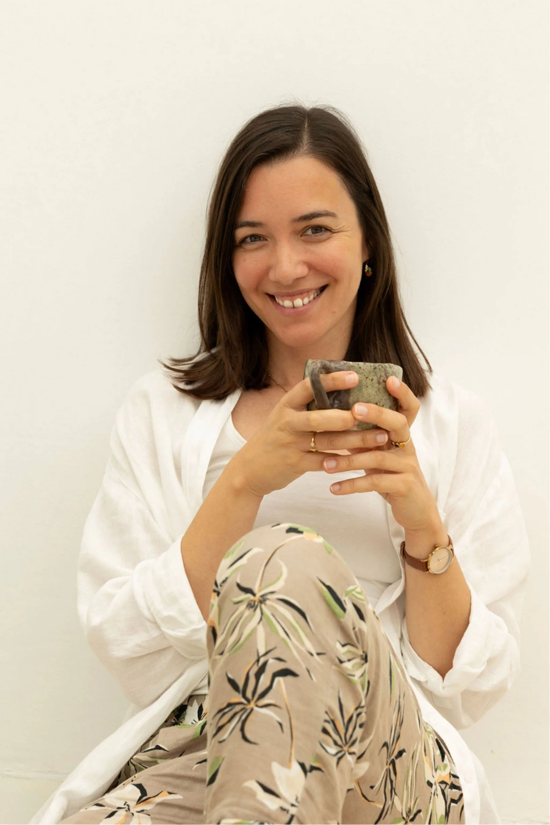 A woman with shoulder-length brown hair smiling, holding a speckled mug, sitting against a plain white wall, wearing a white top and floral patterned pants.