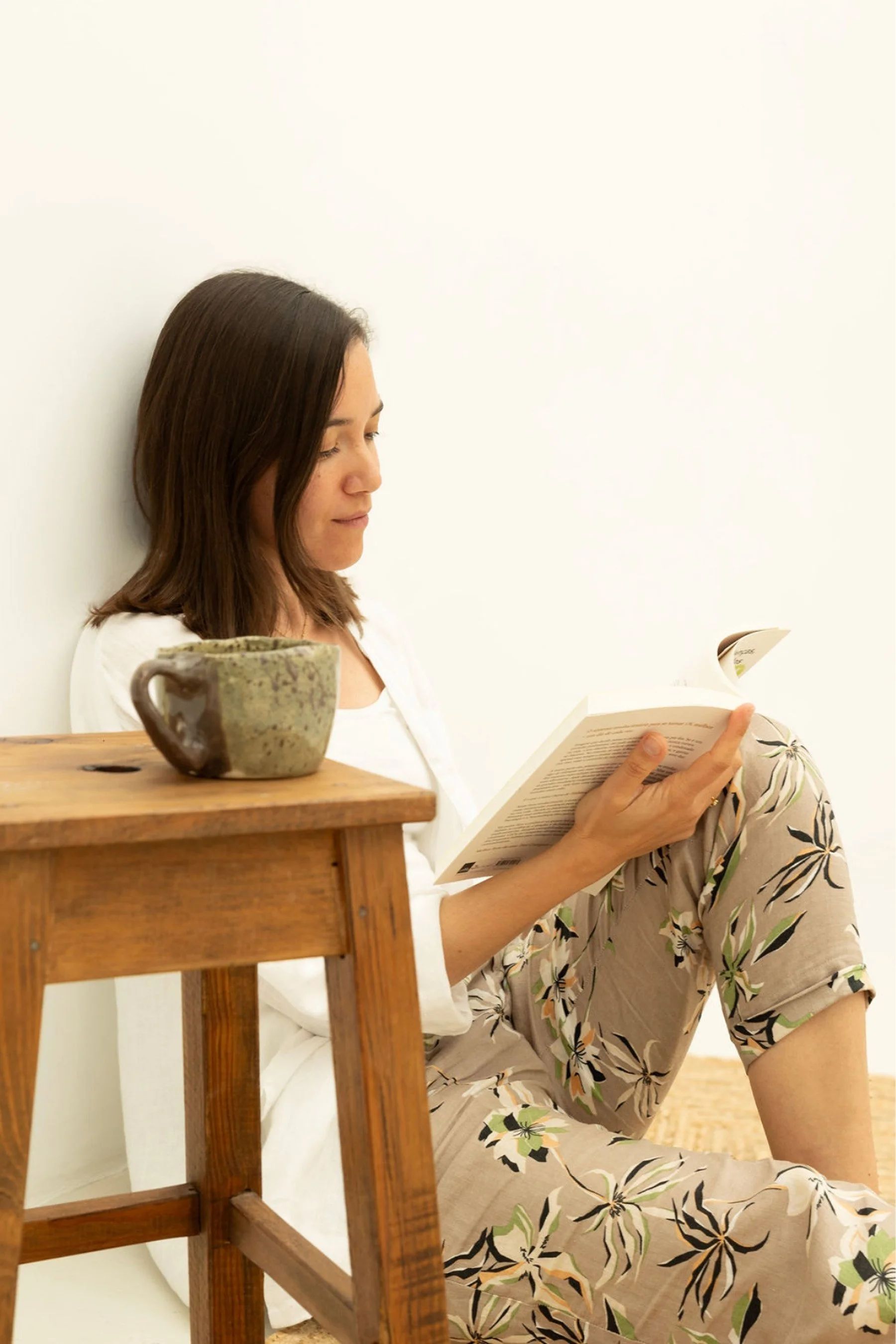 A woman with brown hair is sitting on the floor, leaning against a white wall, reading a book. A ceramic mug is placed on a wooden piece of furniture in front of her. She is wearing a light-colored, floral-patterned dress.