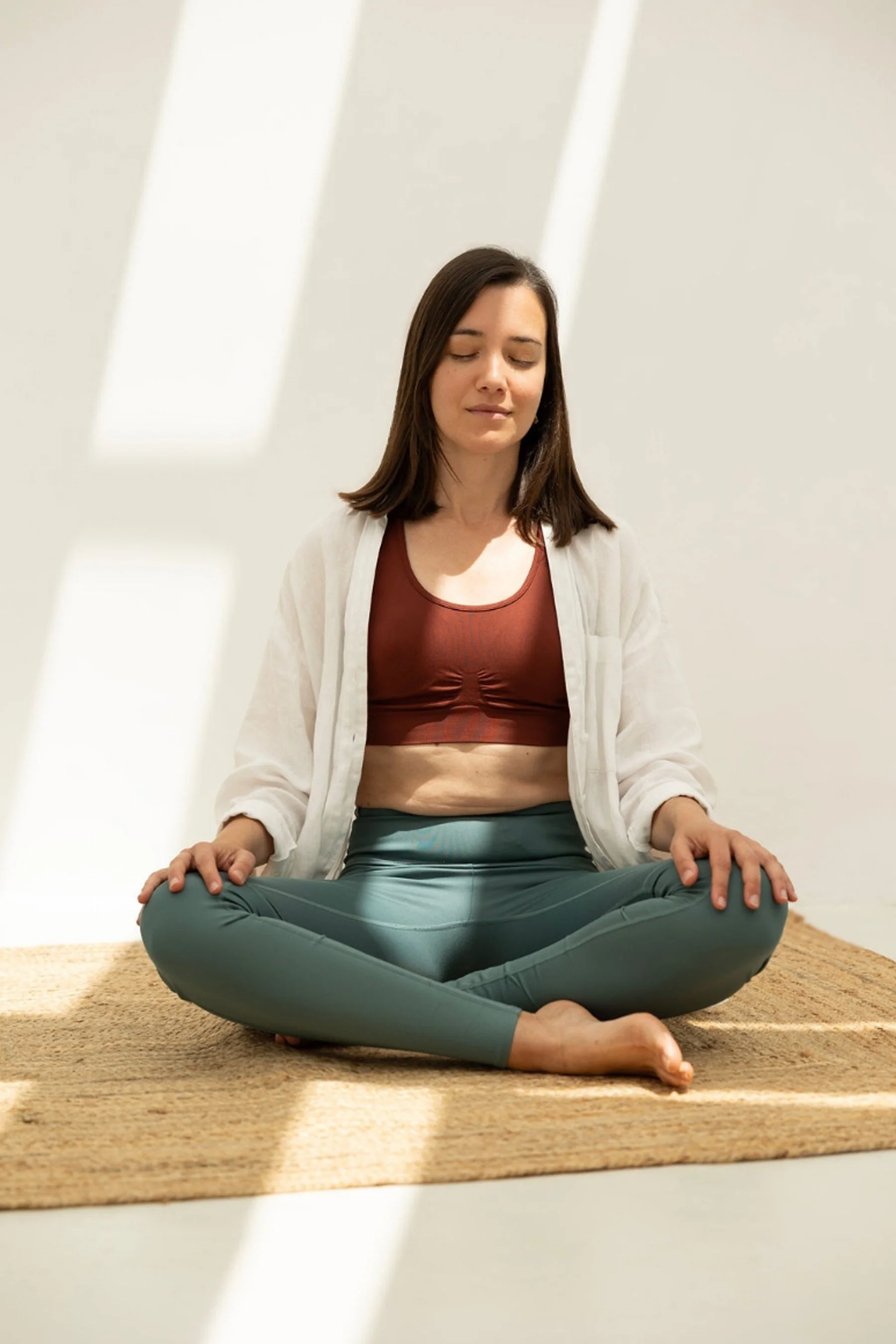 A young woman practicing yoga or meditation indoors on a beige rug, seated cross-legged with her eyes closed, wearing a rust-colored crop top, gray leggings, and a white cardigan, with minimal natural lighting.