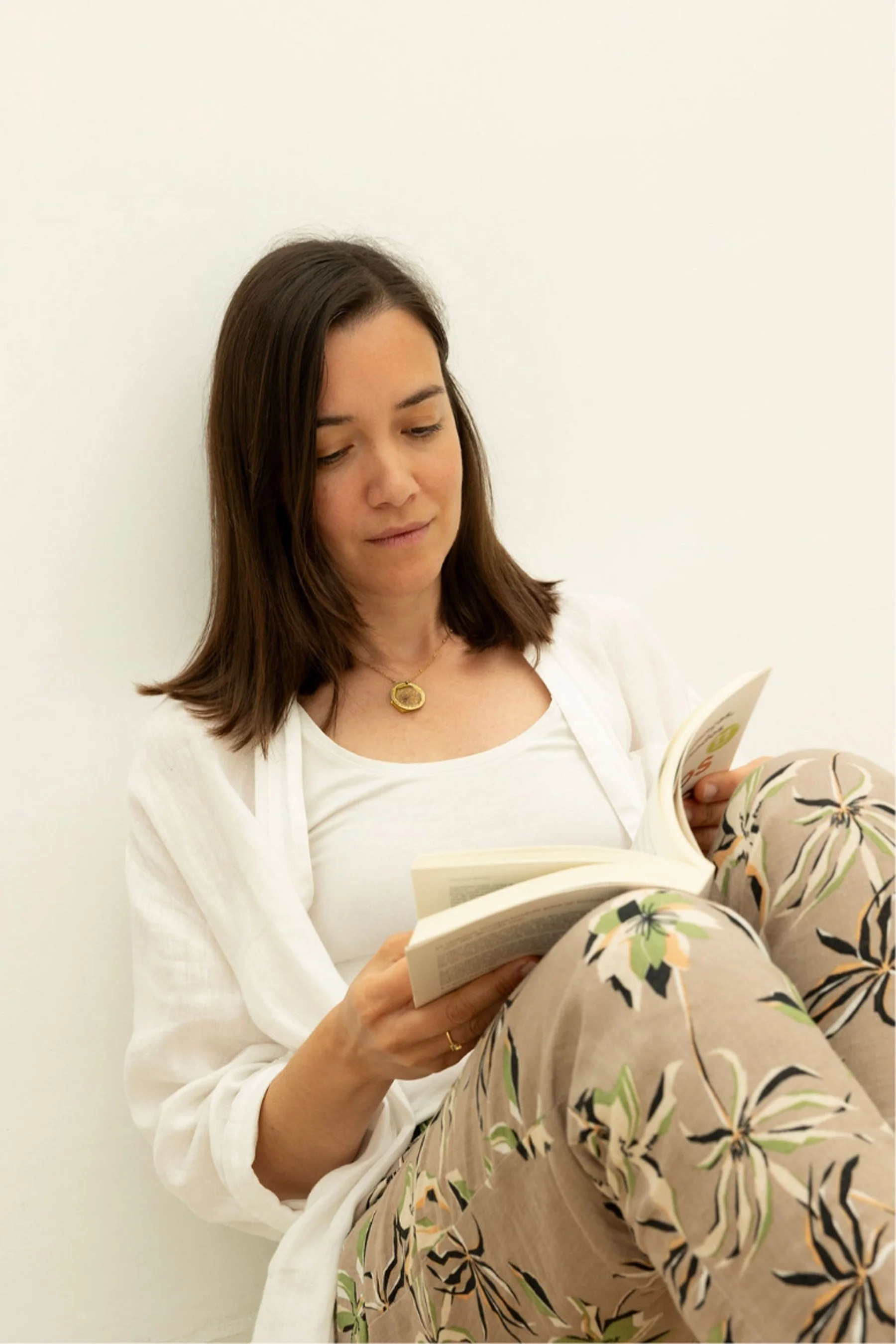 A woman with brown hair reading a book while sitting on the floor against a plain white wall. She is wearing a white top, floral patterned pants, and a gold necklace.