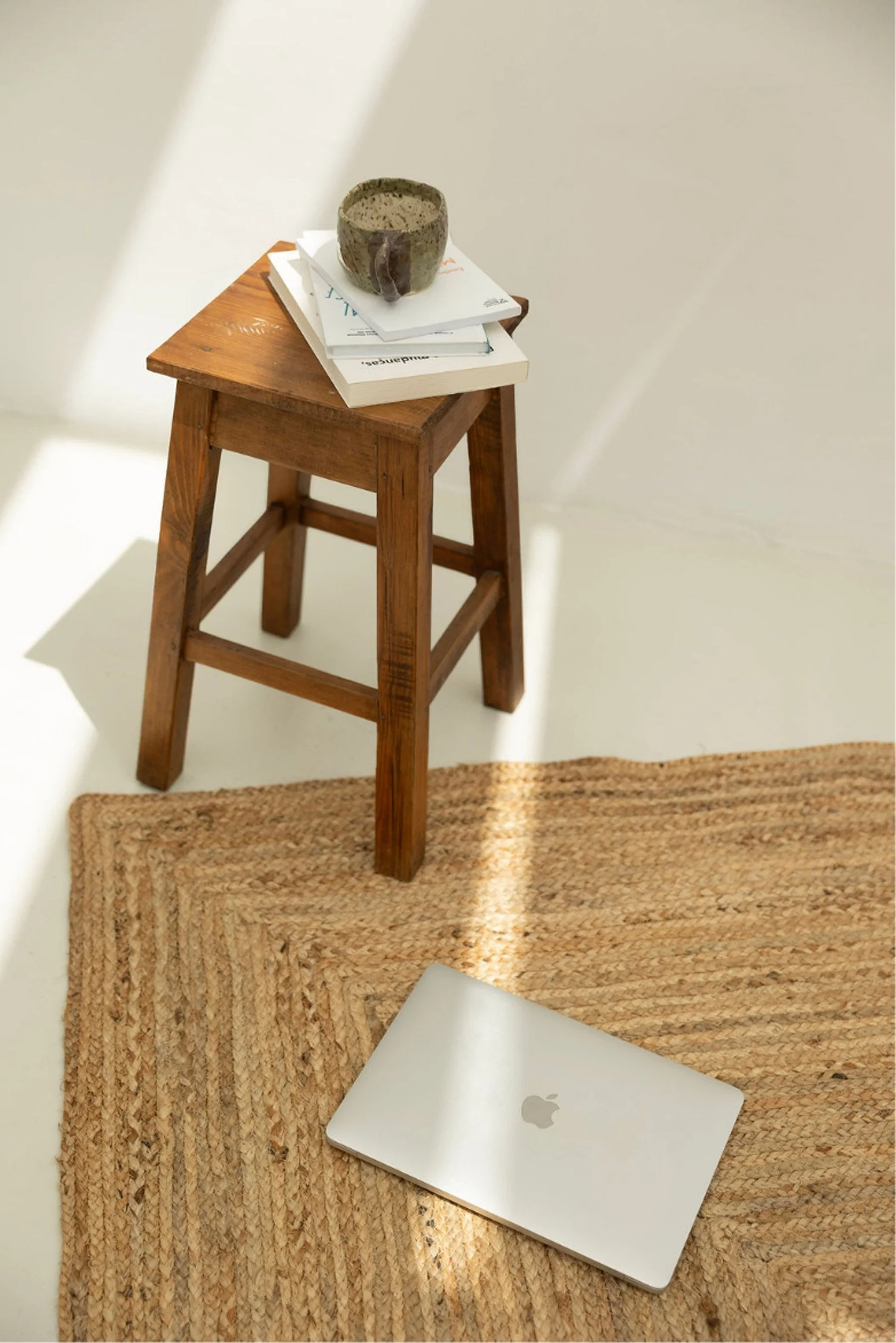 A small wooden stool with a cup and a stack of papers on top, a woven rug, and a closed silver Apple MacBook on the rug, against a plain beige wall.
