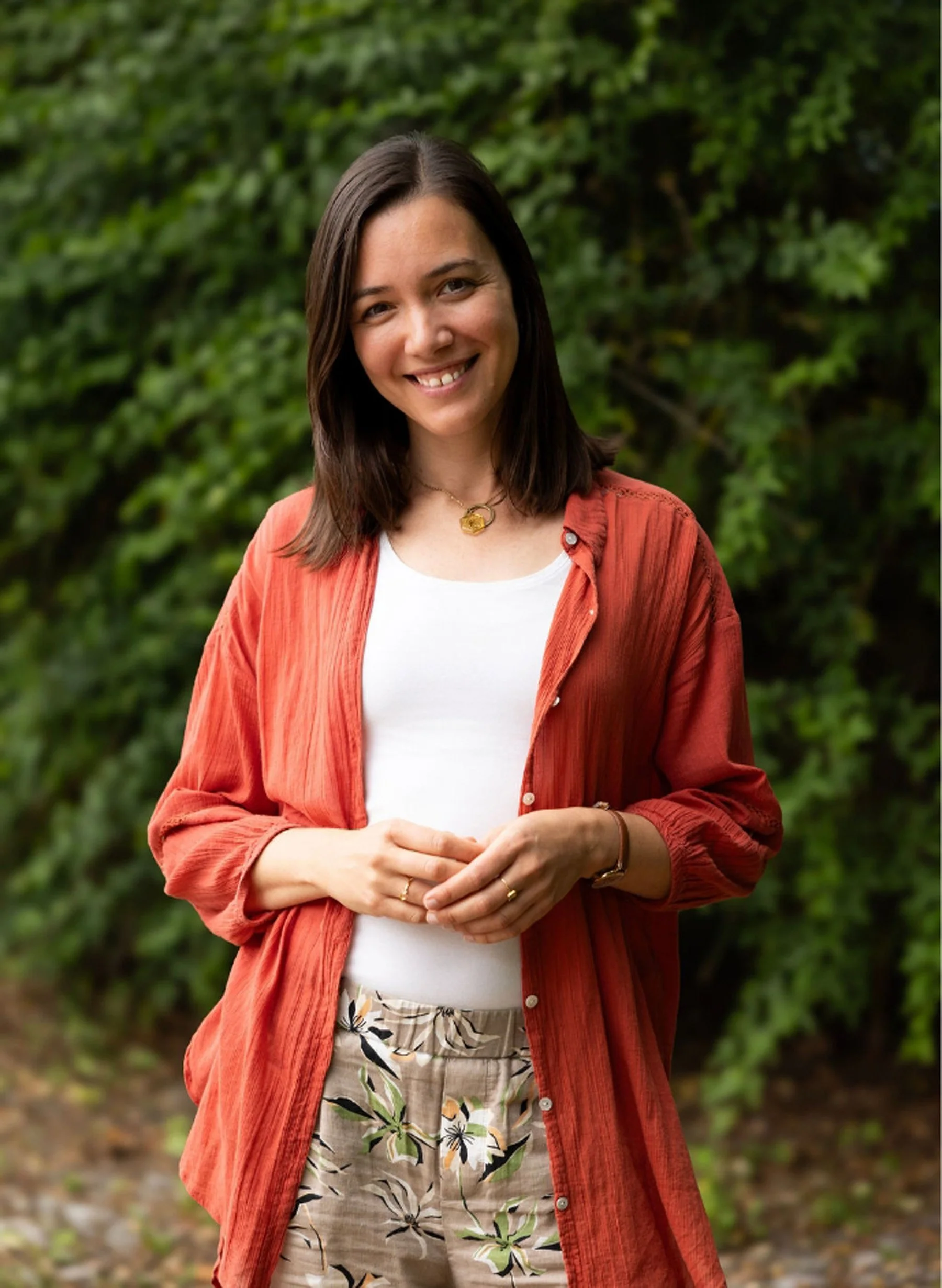 A smiling woman with shoulder-length brown hair wearing a white top, orange-red button-up shirt, patterned shorts, and a gold necklace standing outdoors with green foliage in the background.