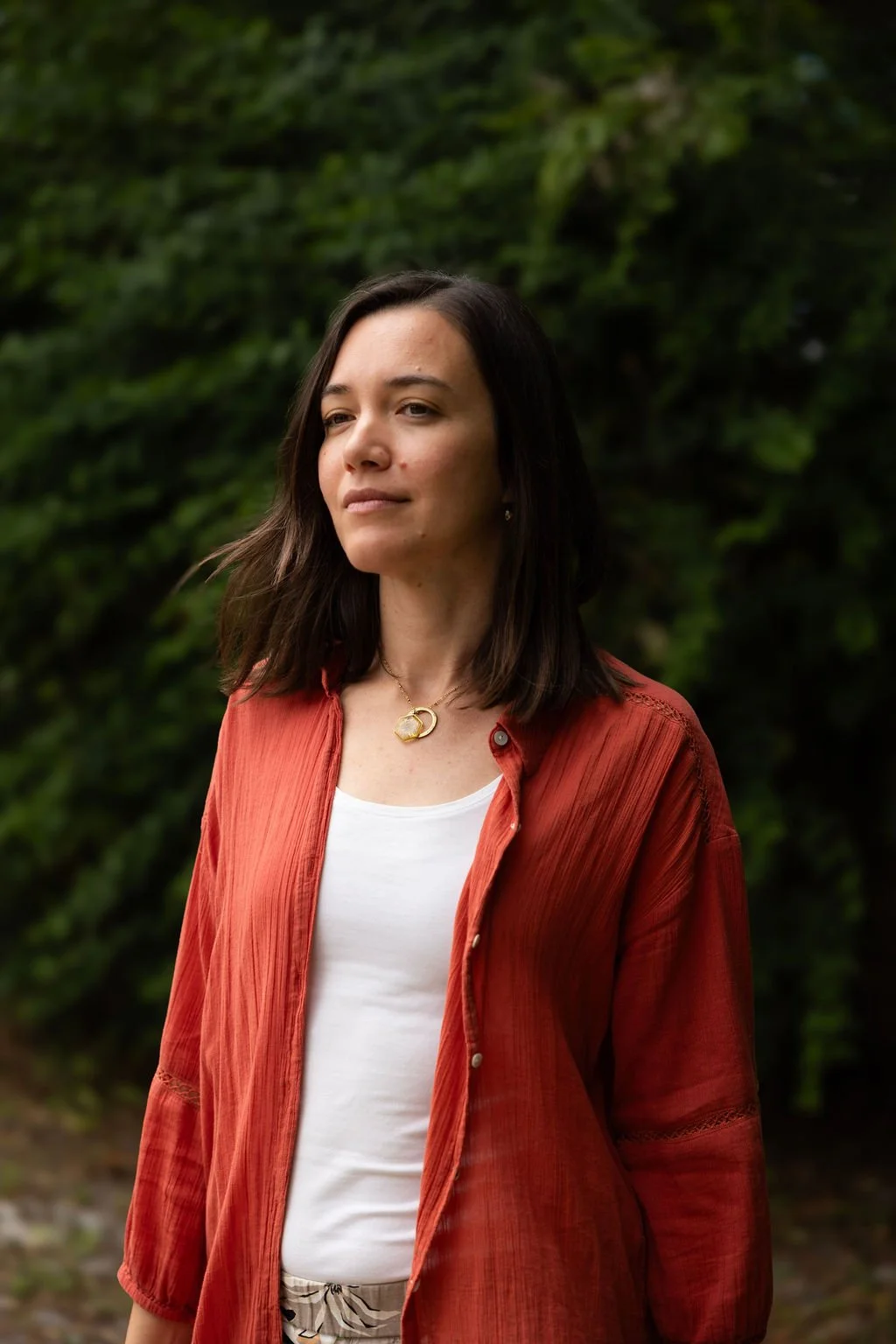 A woman with dark brown hair wearing a white top, a red-orange shirt, and a gold necklace, standing outdoors with green foliage in the background.