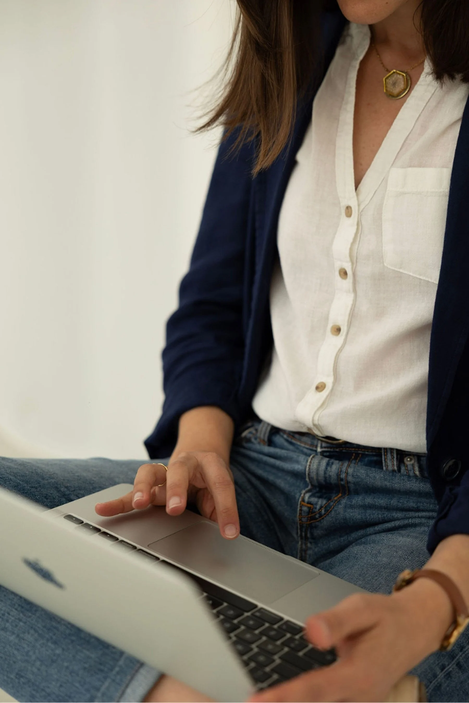 A woman using a laptop computer while working or browsing, dressed in casual jeans and a white button-up shirt with a navy jacket, with jewelry including a gold necklace and ring, in a well-lit indoor setting.