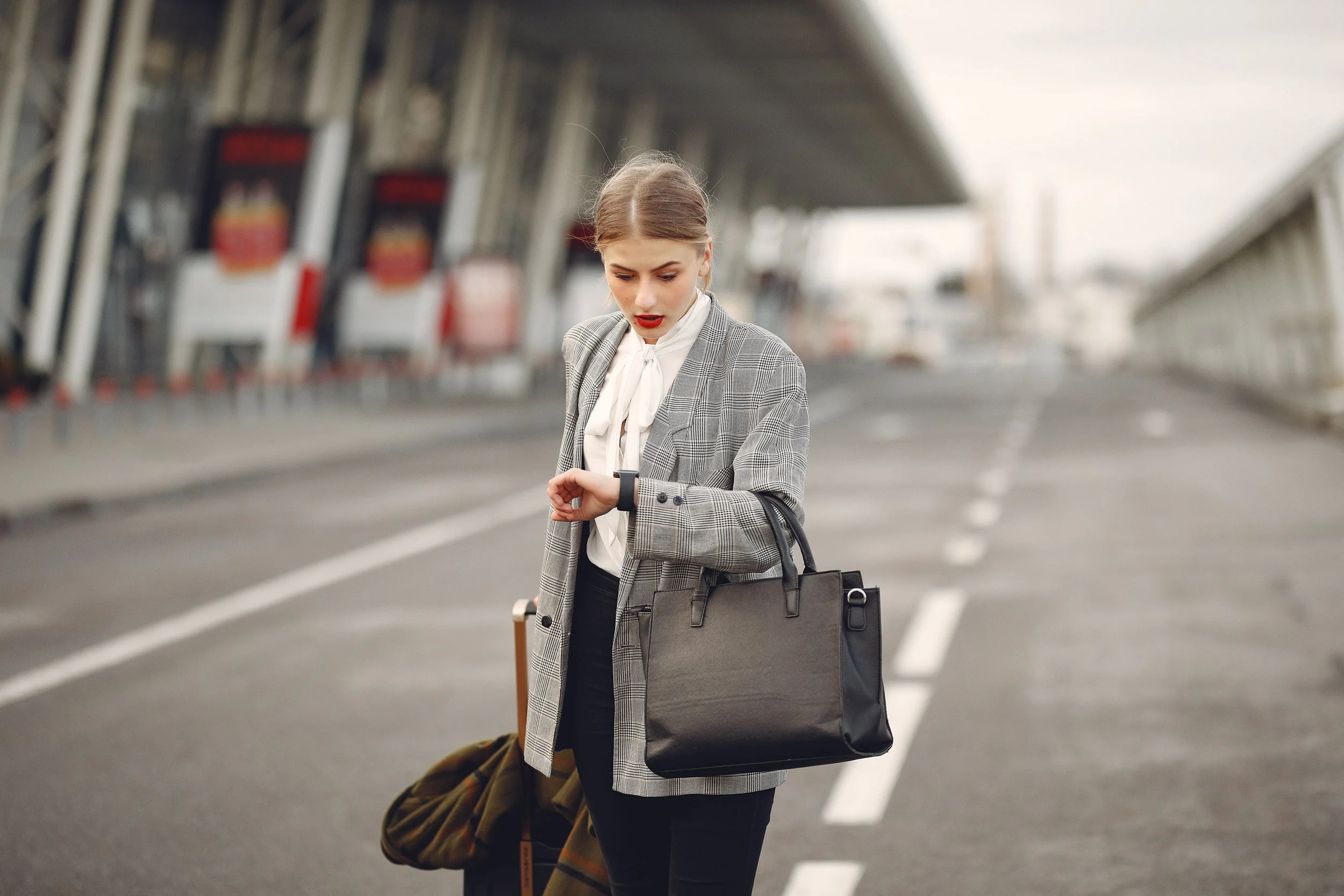 A woman with light brown hair, dressed in a gray plaid blazer and white blouse, stands on an empty road with luggage and a bag, looking at her smartwatch.