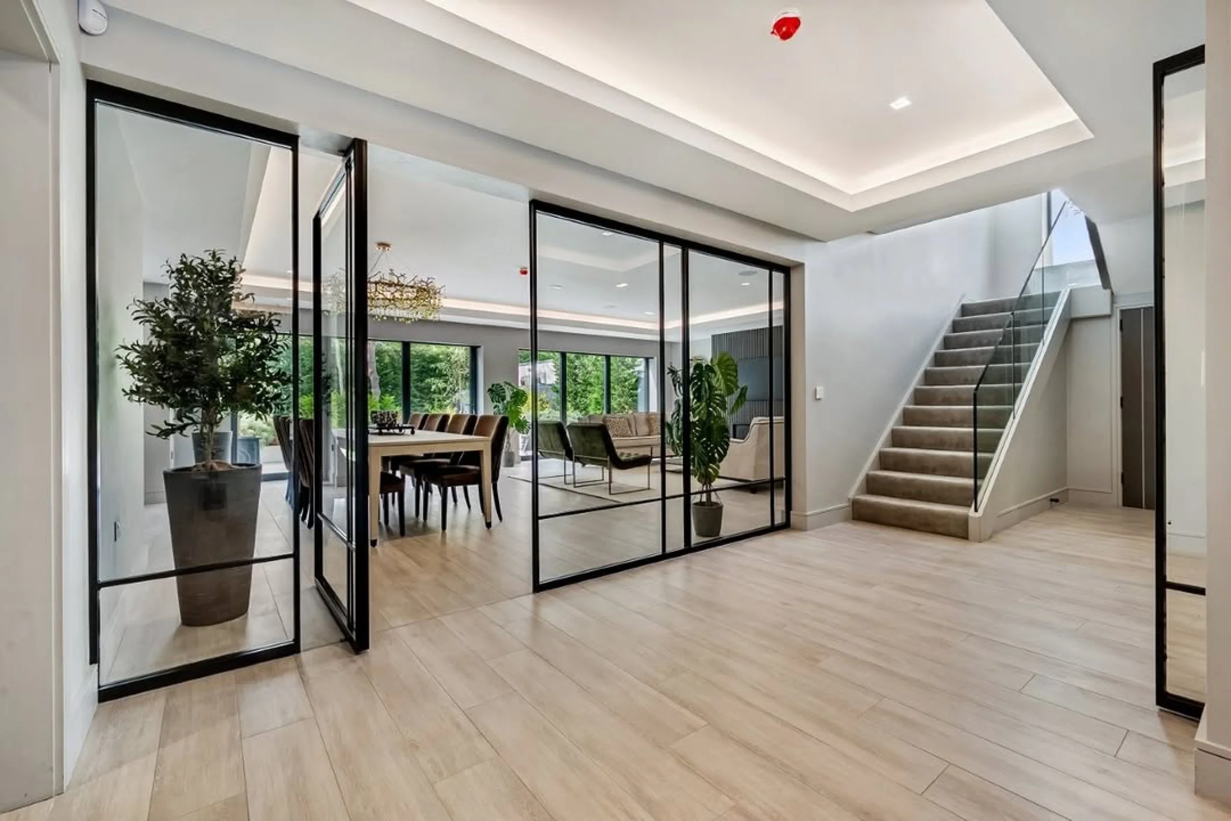 Interior view of a modern house with a staircase, glass partition with black frame, a dining area with a large table, chairs, and potted plants, large windows showing outdoor greenery, and a ceiling with recessed lighting.