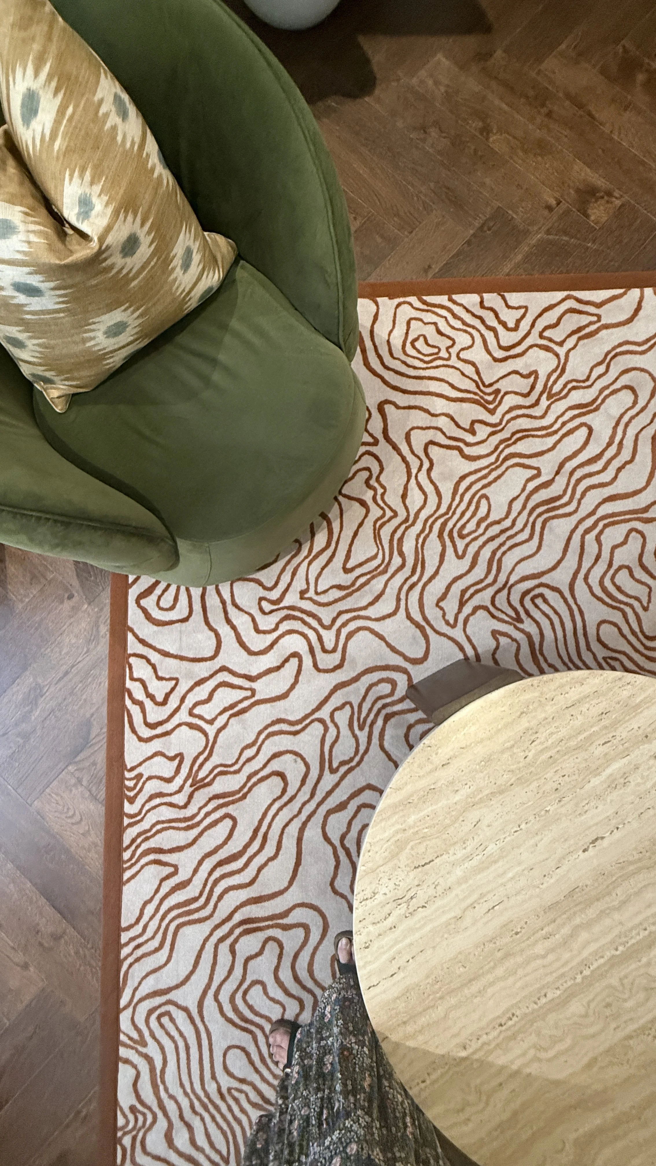 View of a patterned area rug on a wooden floor, a green upholstered chair with a decorative pillow, and part of a round table with a beige surface, with a person wearing a patterned skirt and sandals standing nearby.