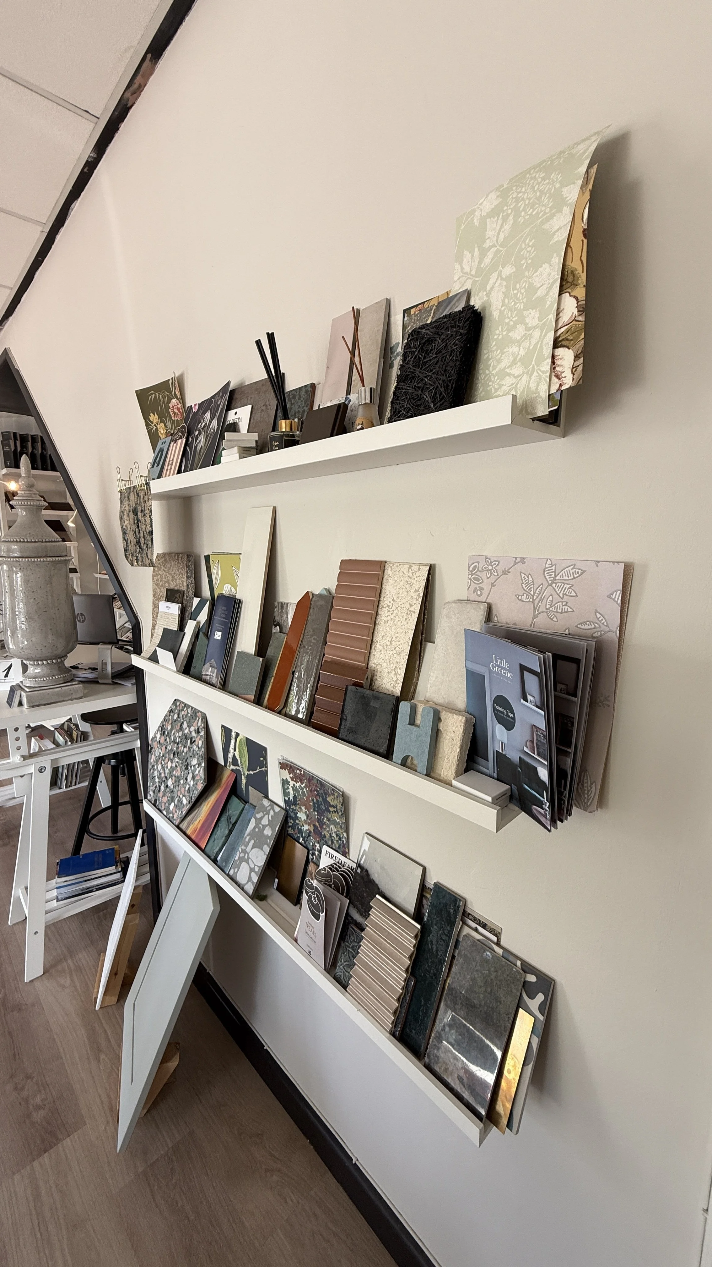 Interior view of a display wall with various sample tiles and fabric swatches arranged on three white shelves in a design or home improvement store.