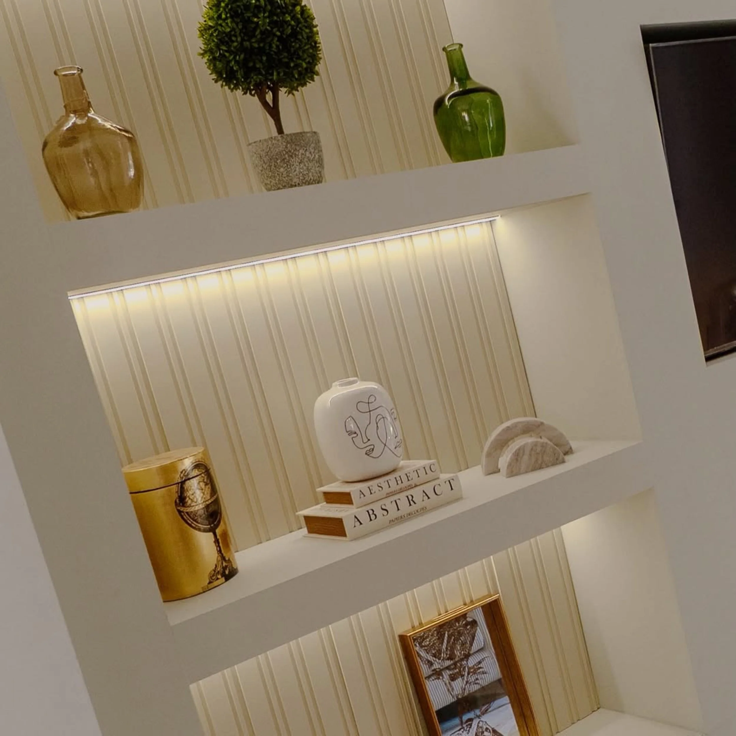 A wall shelf decorated with a potted topiary tree, two decorative vases (one green, one gold), and several books with titles related to aesthetics and abstract art. The shelf is under a ceiling light fixture with horizontal blinds below.