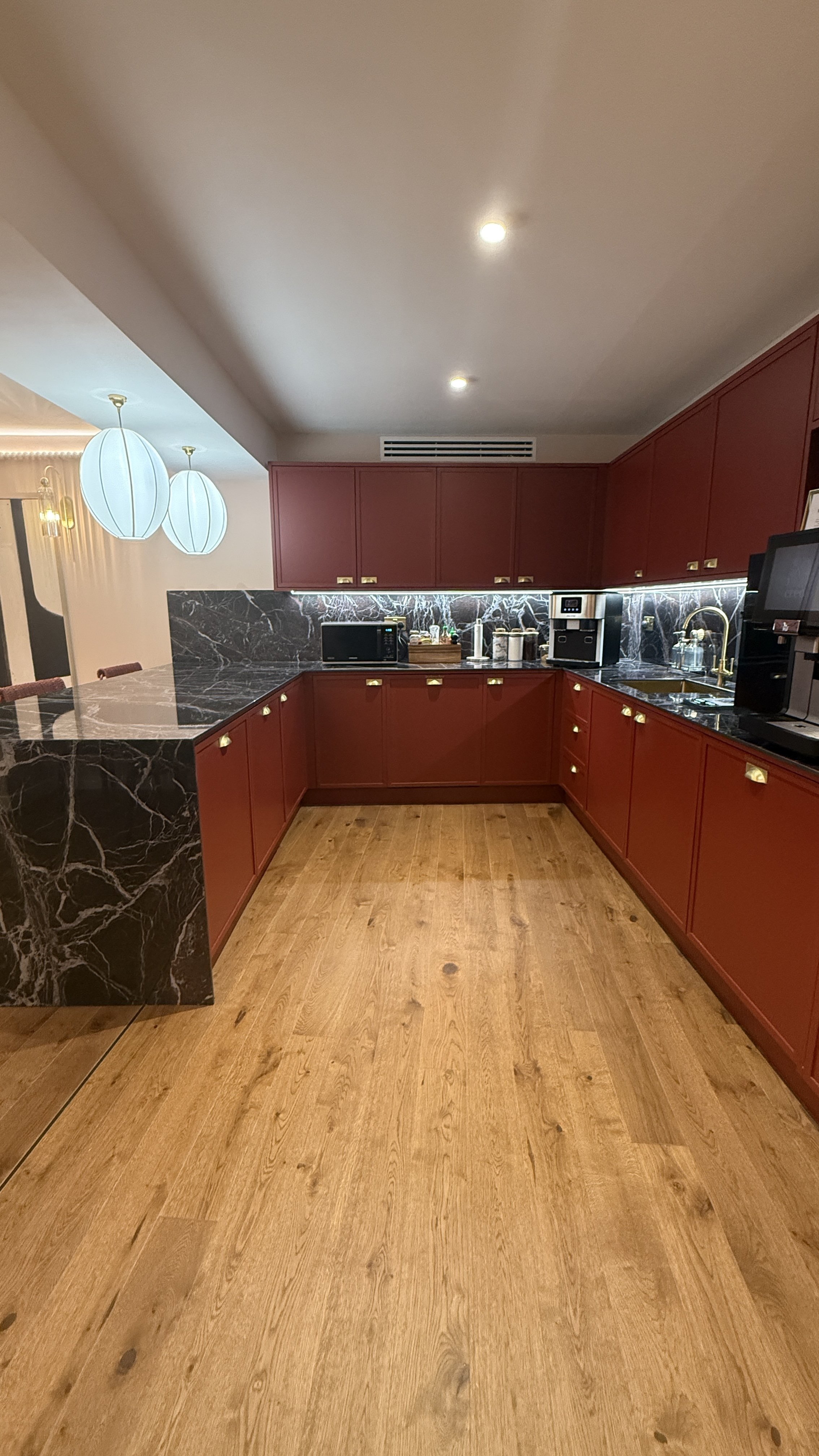 A kitchen with red cabinets, black marble countertops, wooden flooring, and modern appliances including a microwave and coffee machine.