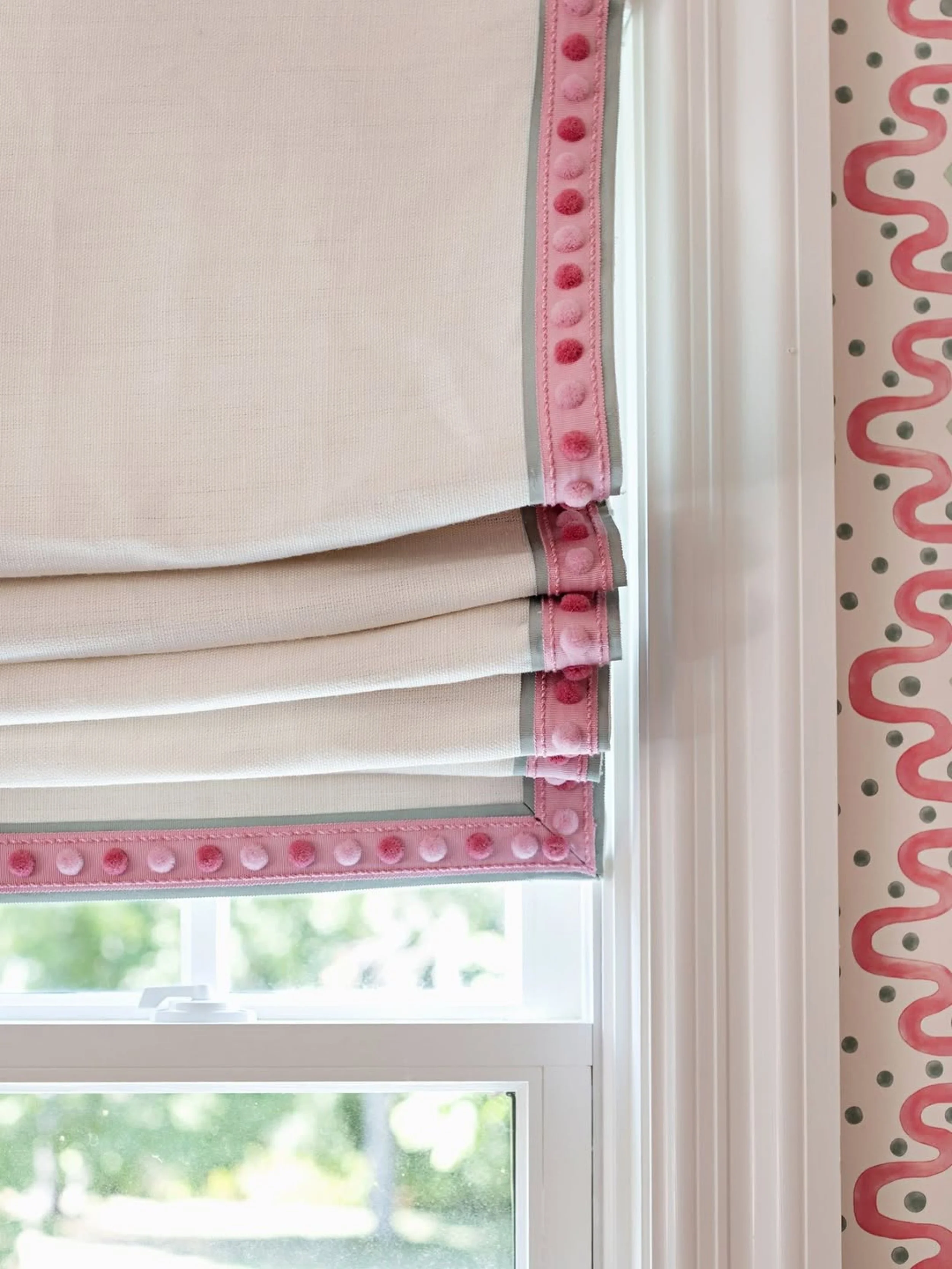 A white window with a pink curtain that has a pink pom-pom trim and pink and red polka dots, showing a green outdoor scene with trees and grass.