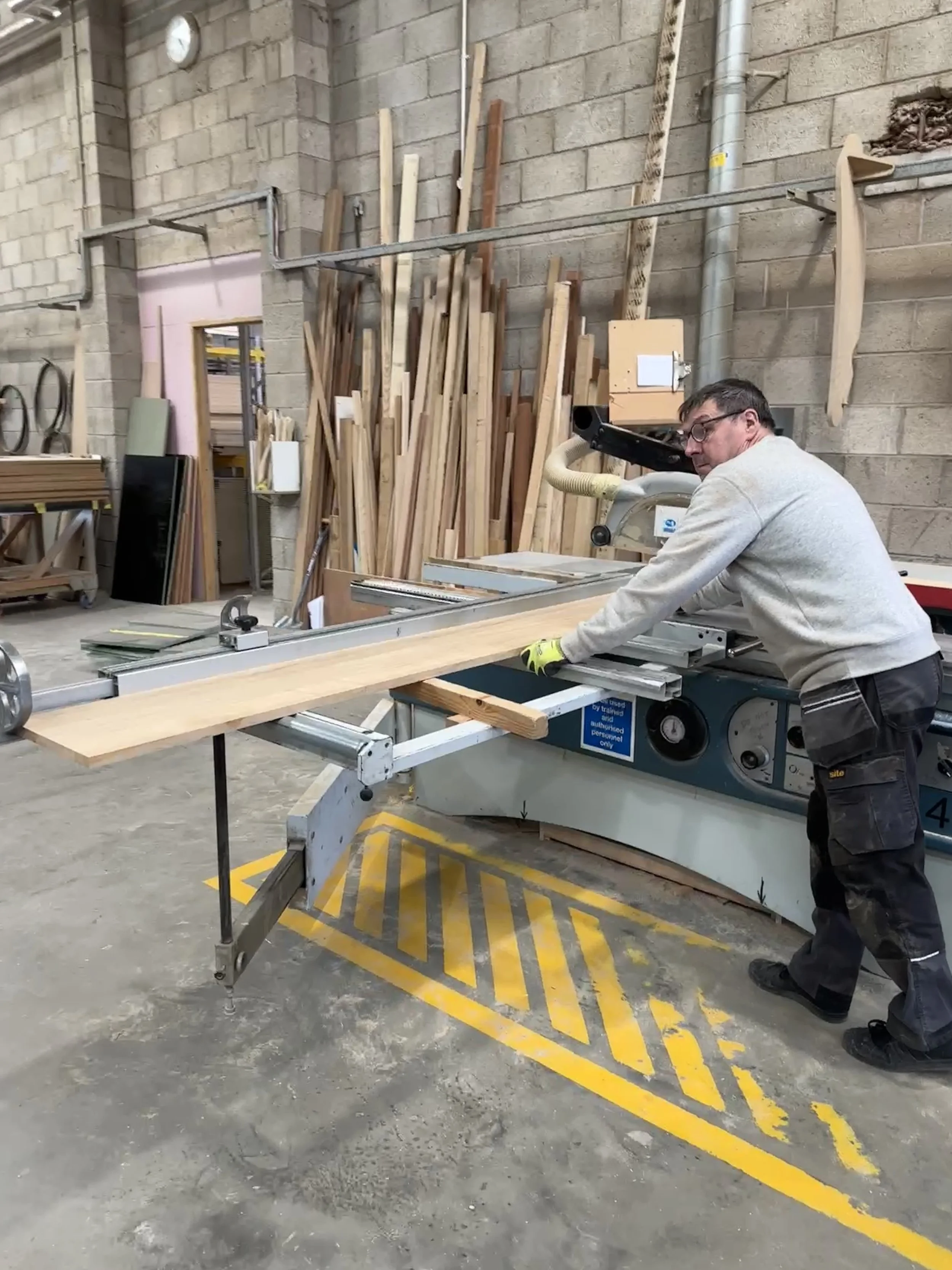 Man operating a sliding table saw in a woodworking shop with wood planks in the background.