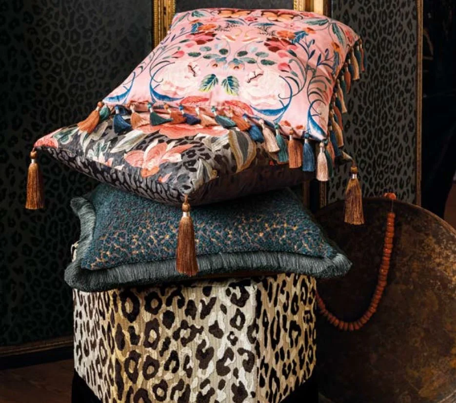 A stack of four decorative pillows on a leopard print stool. The top pillow has a pink floral design with tassels, the second pillow is dark with a textured pattern, the third pillow is velvet with small gold flecks, and the bottom pillow features a black and cream leopard print. Behind the pillows, there is a black wall with a subtle animal print pattern.
