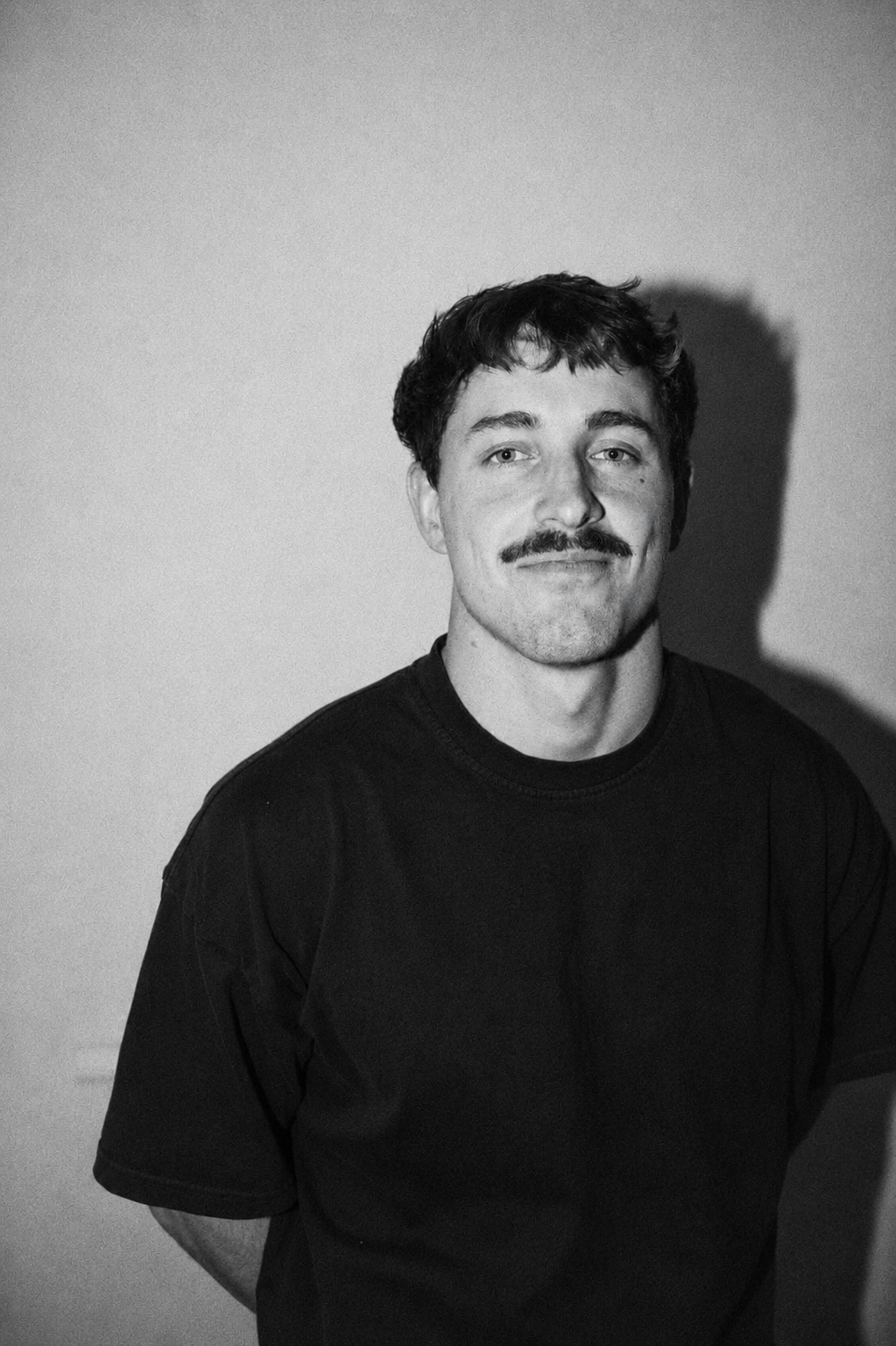 Black and white portrait of a young man with short wavy hair and a mustache, wearing a plain dark t-shirt, standing against a plain background with a shadow cast on the wall.