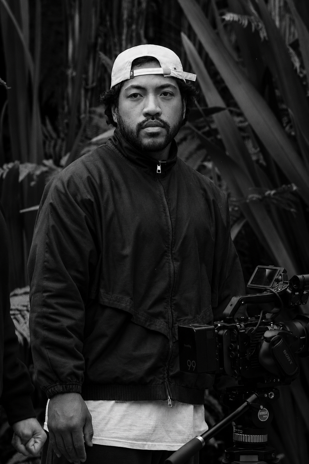 A man with a beard and mustache wearing a backwards baseball cap and a black jacket is standing outdoors in front of tall plants, possibly crops, with a professional video camera on a tripod in front of him.