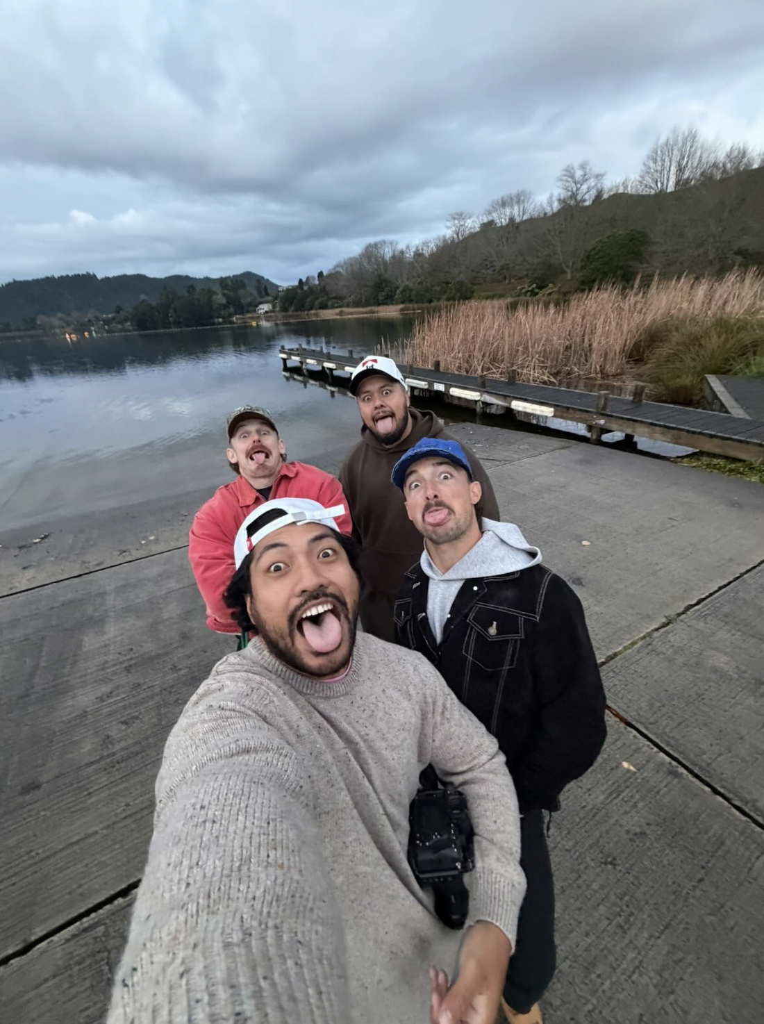 Four men taking a selfie during daytime by a lake with a dock, hills, and cloudy sky in the background, all sticking out their tongues.