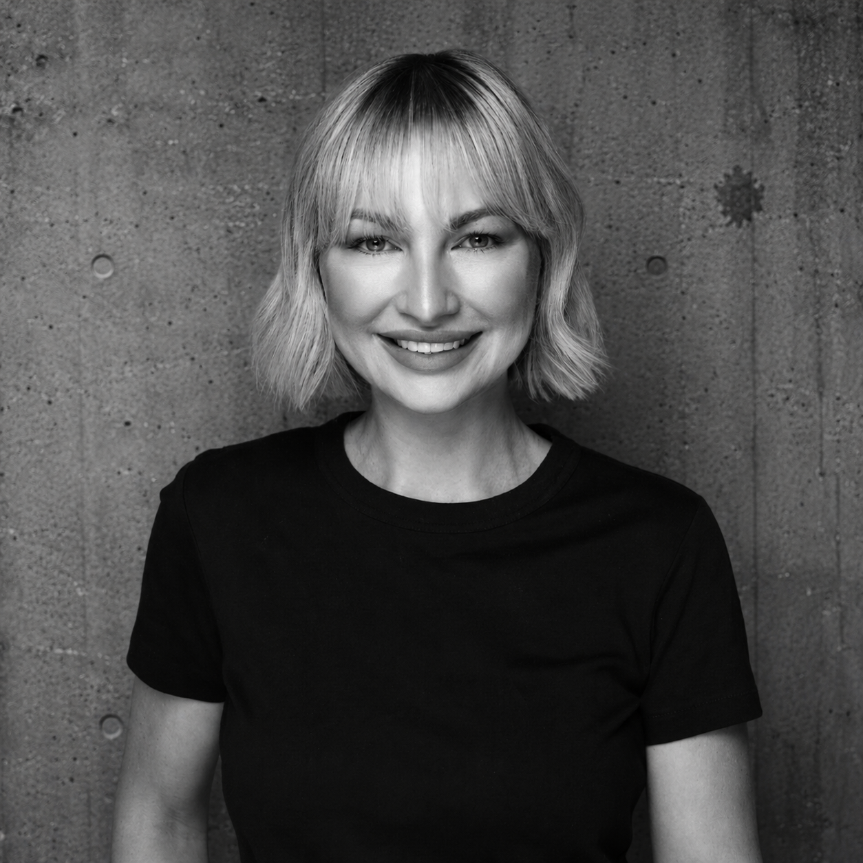Black and white portrait of a woman with blonde, shoulder-length hair, smiling, wearing a black t-shirt, standing against a textured concrete background.