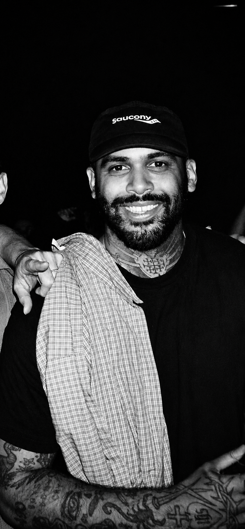 A smiling man with a beard and tattoos, wearing a Saucony cap and a black t-shirt, holding a checkered shirt over his shoulder in a dark environment.