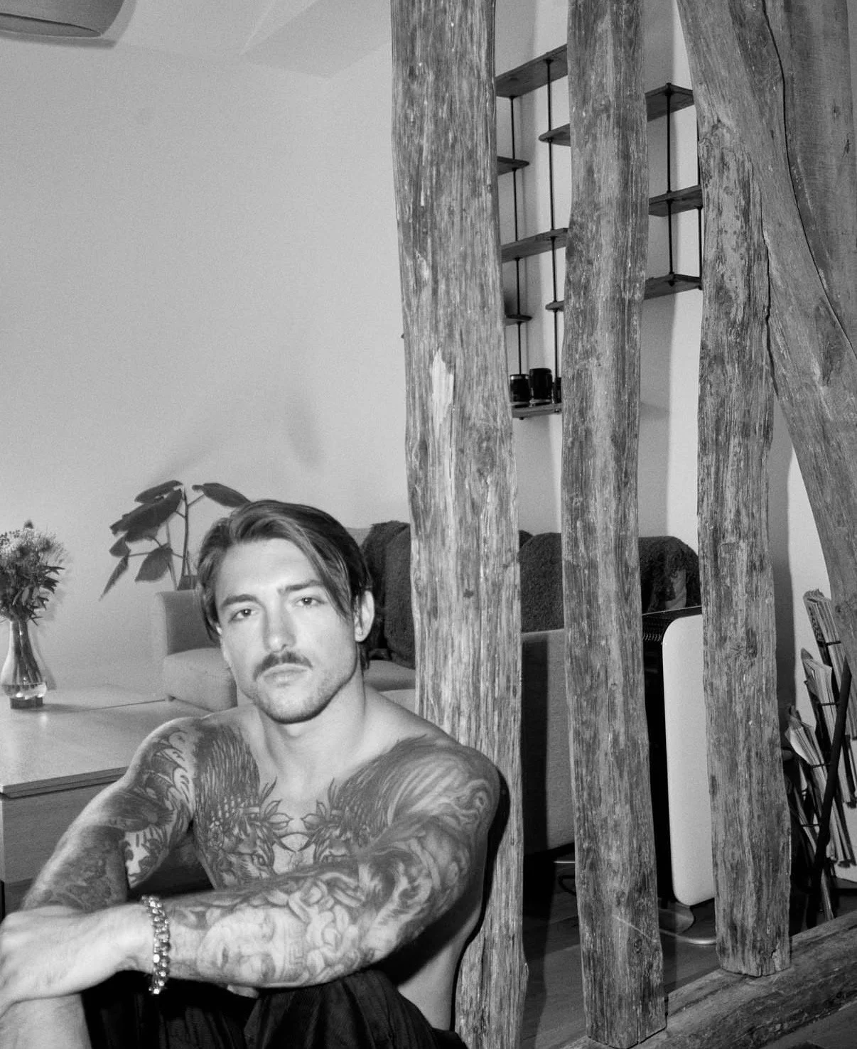A young man with tattoos on his arms and chest sitting in a living room, with wooden beams and a shelf with small black objects in the background. Black and white photo.