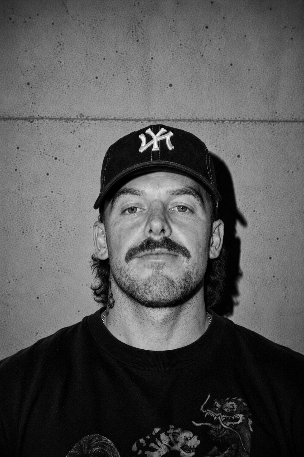 Black and white portrait of a man with a moustache and beard wearing a baseball cap with the New York Yankees logo, standing against a textured wall.