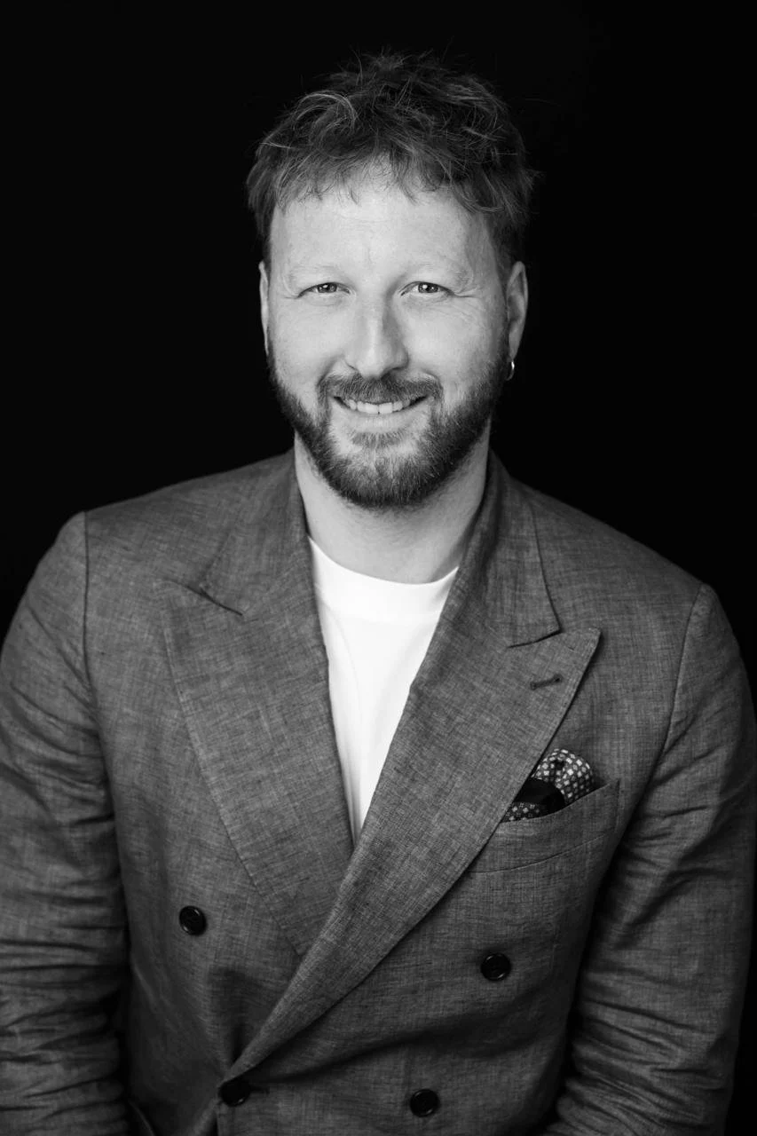 Black and white portrait of a smiling man with a beard, wearing a double-breasted blazer, a white t-shirt, and a pocket square, against a dark background.