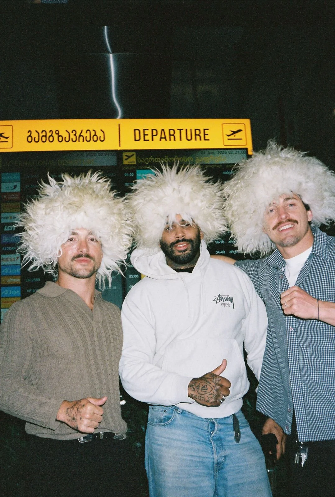 Three men standing in an airport terminal under a 'Departure' sign, wearing large, fluffy white hats.