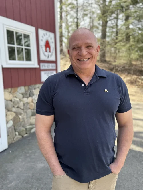 A smiling man in a navy blue polo shirt standing outside near a red building with a stone foundation and a sign that says "The Red Barn" in a wooded area.