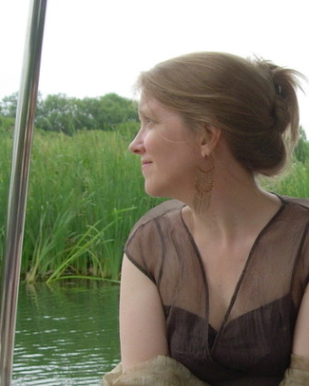 Emily Van Evera sits with light brown hair tied back, wearing earrings and a sheer, brown top, looking to the side while on a boat surrounded by green water and tall grass.