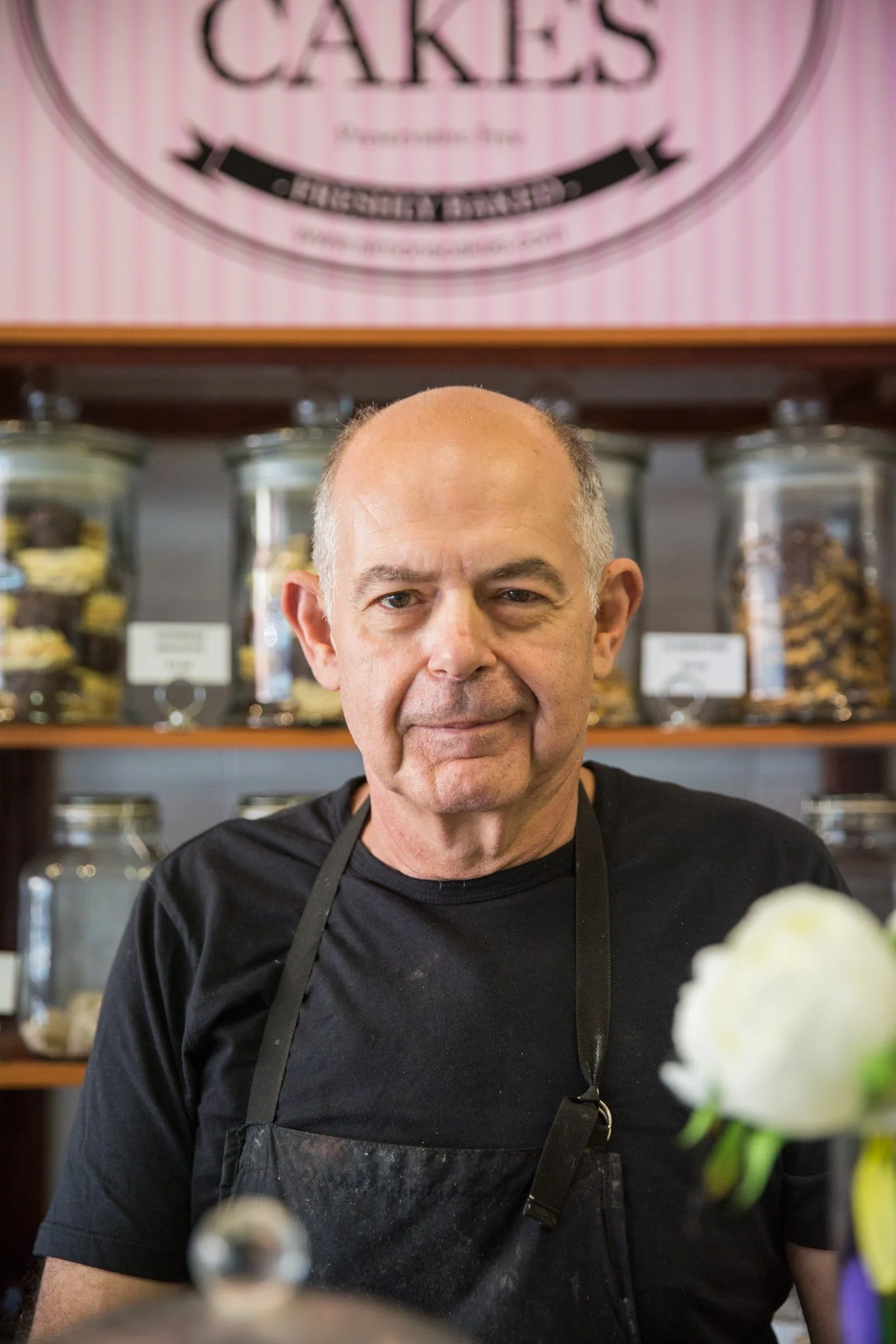A middle-aged man with a bald head and gray hair on the sides, wearing a black shirt and apron, is standing in front of a display of jars filled with baked goods and candies in a shop or bakery. There is a pink and white striped sign above him that says 'Cakes'.