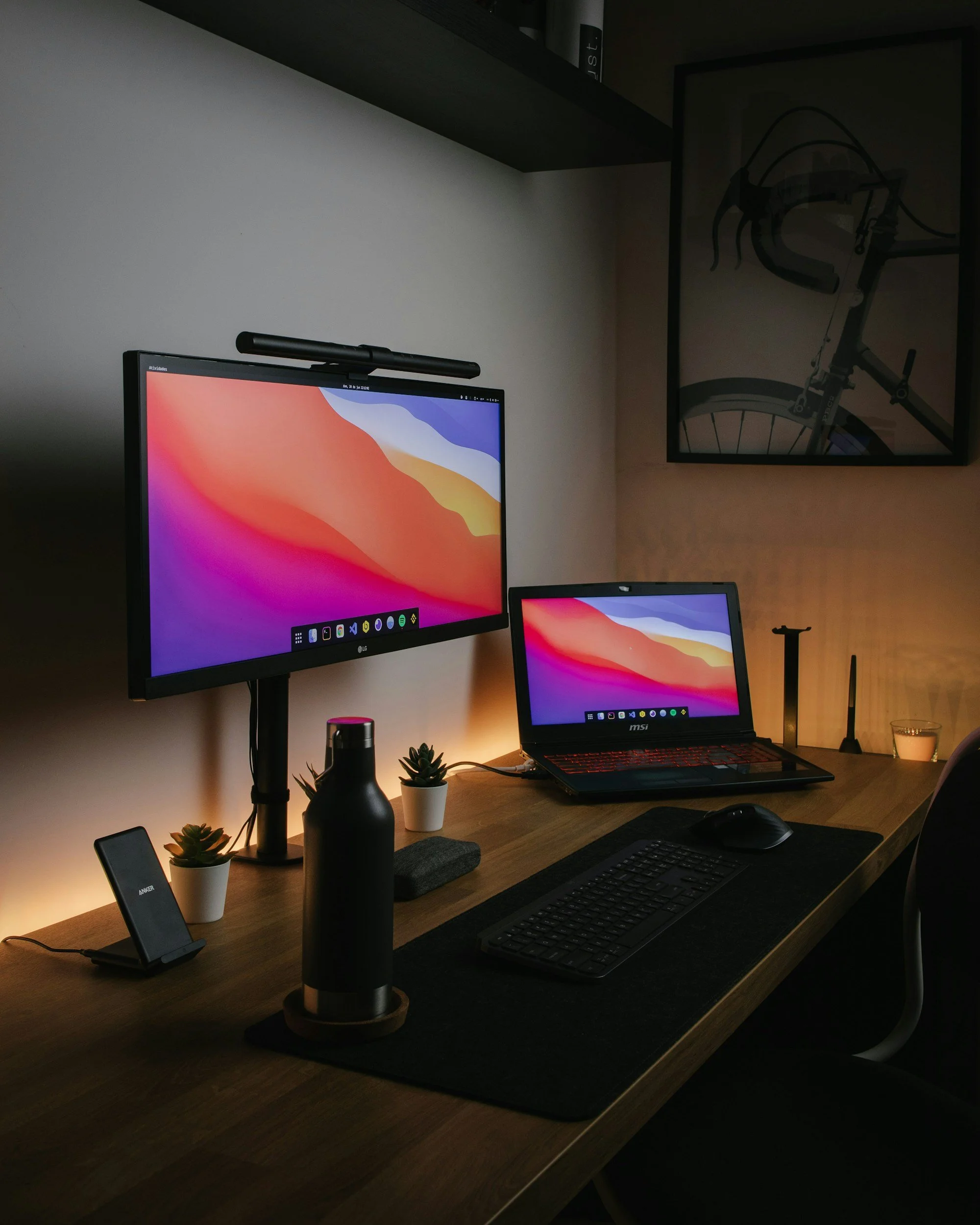 A computer workspace with two monitors displaying colorful backgrounds, a laptop, a keyboard, a mouse, a smartphone on a stand, a water bottle, two small potted succulents, a candle, and a framed picture of a bicycle on the wall.