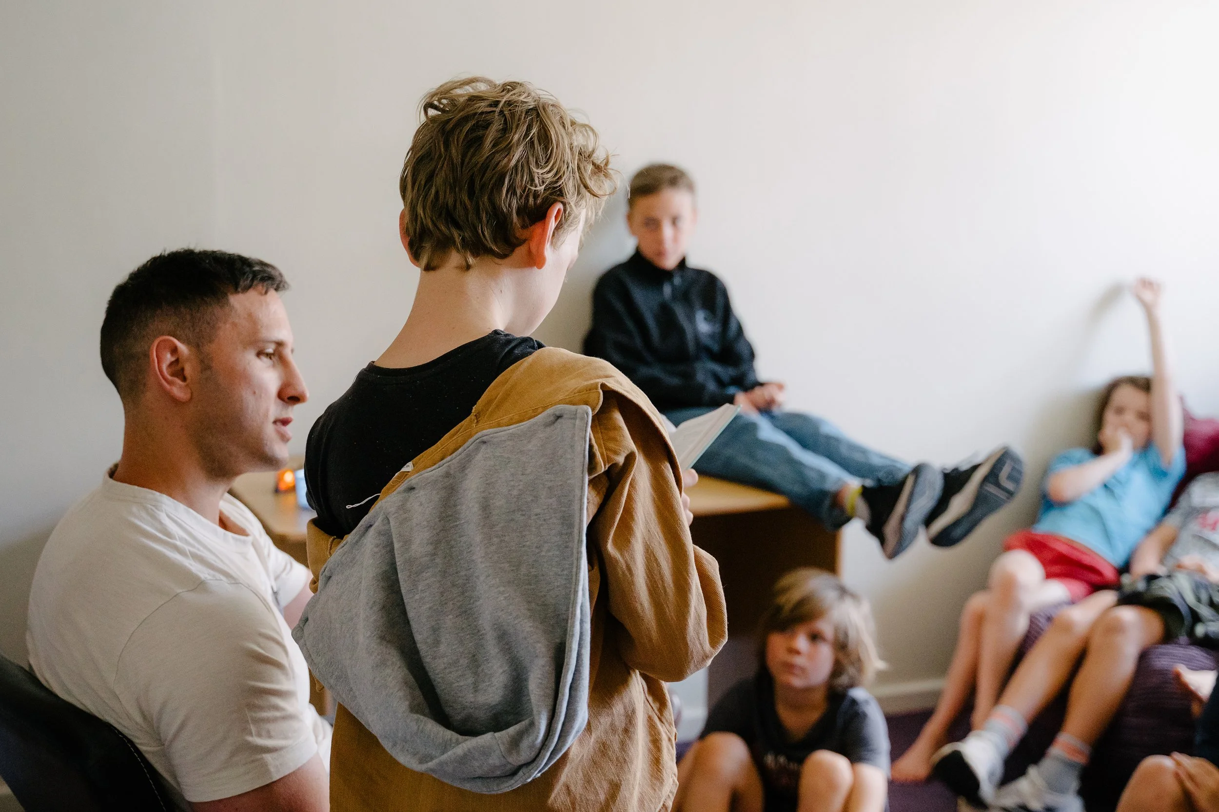 A group of children and teenagers in a classroom, with one girl standing and reading, and others sitting against a wall, some raising their hands.