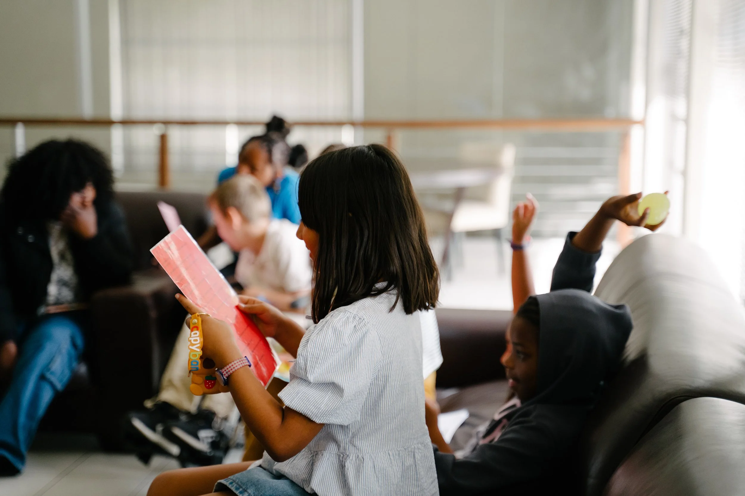 Children sitting in a waiting area, some reading, one holding an apple, in an indoor space with natural light.