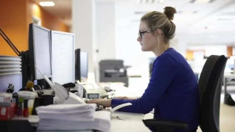 En kvinne med briller jobber på kontor, sitter foran en datamaskin med flere skjermer.