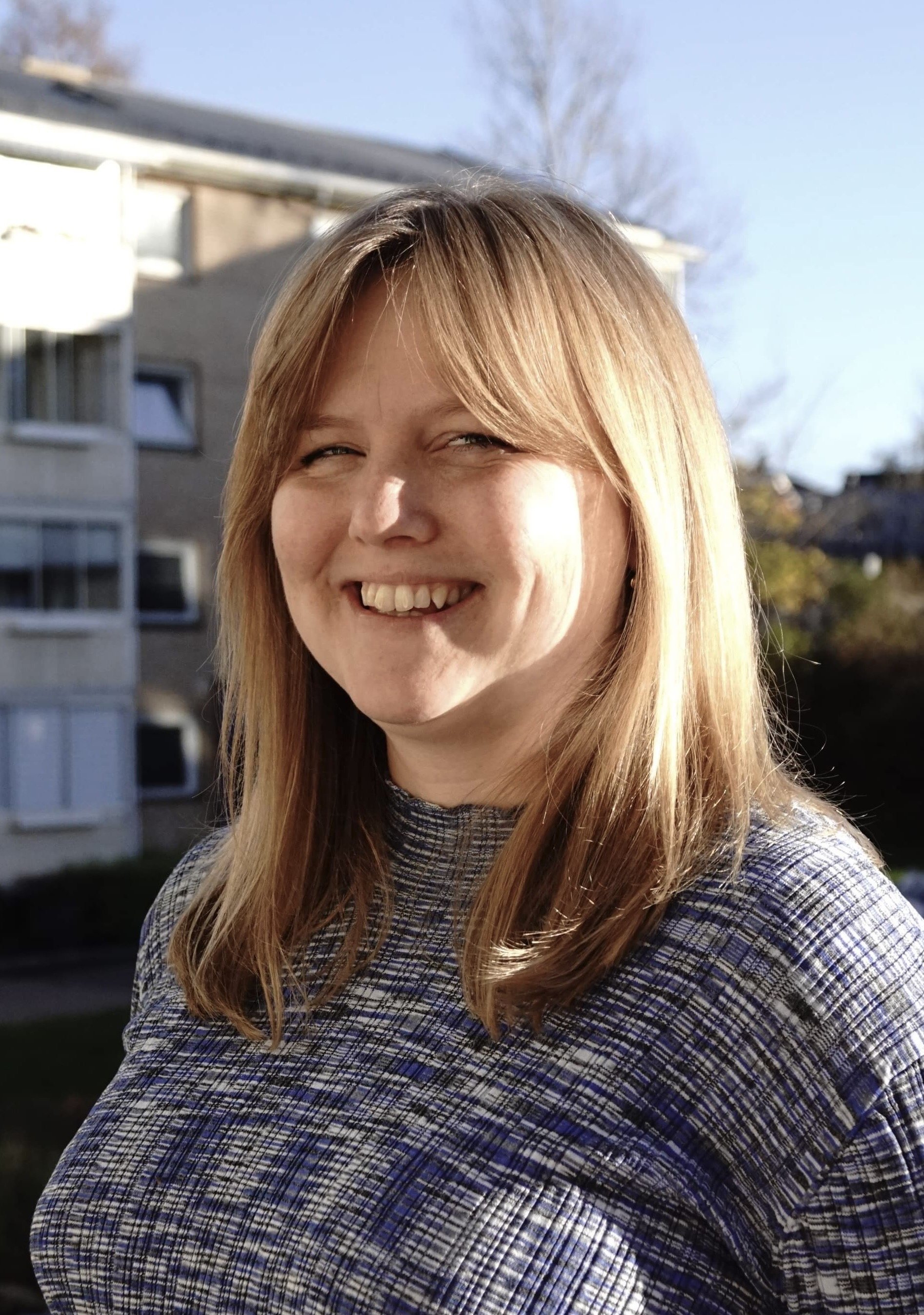 Et smilende kvinne med lang, blond hår, kledd i en rutete skjorte, ute i solen med en bygning i bakgrunnen.