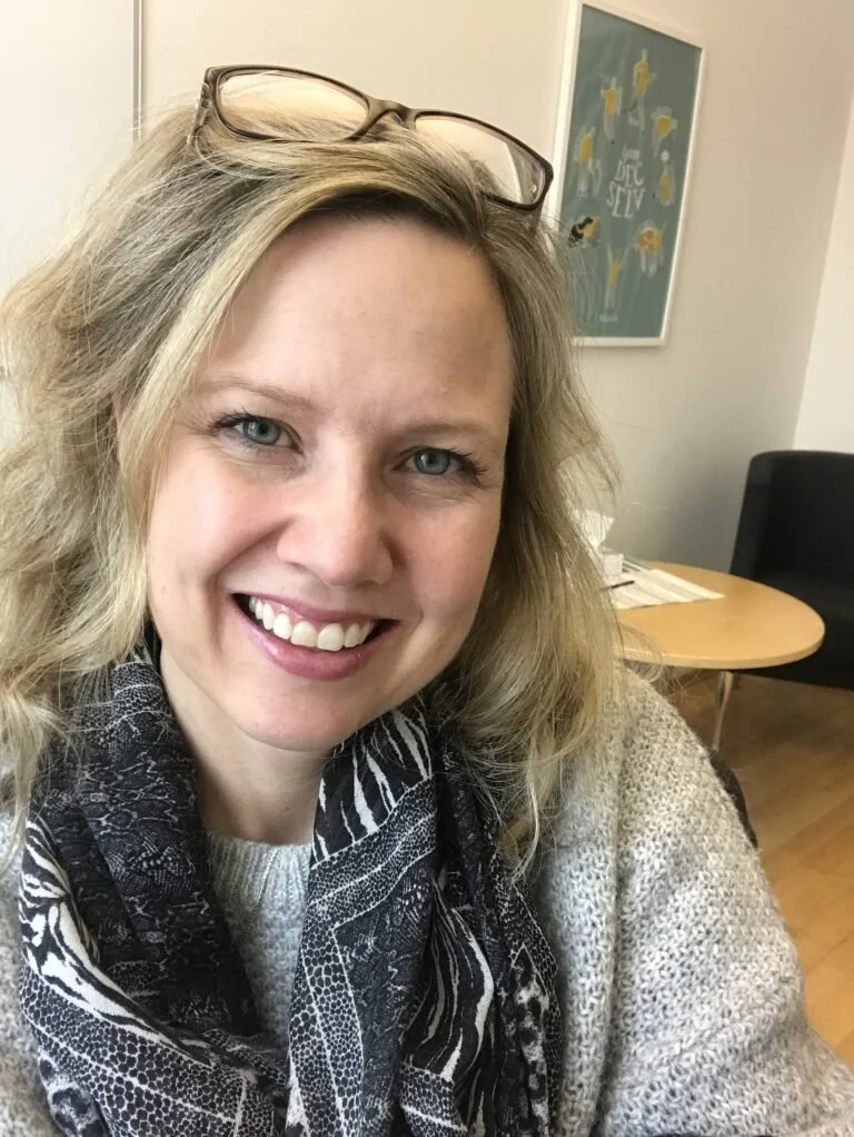 En smilende kvinne med blond, krøllete hår og briller på hodet. Hun har på seg en svart og hvit snusbane og en lys genser. Bak henne er det et skrivebord og en vegg med kunstverk.