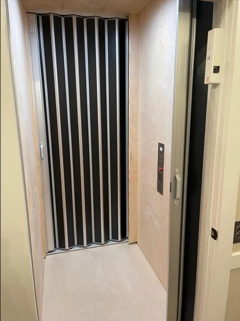 Inside an elevator with folding accordion doors, beige walls, and elevator control buttons on the right side.